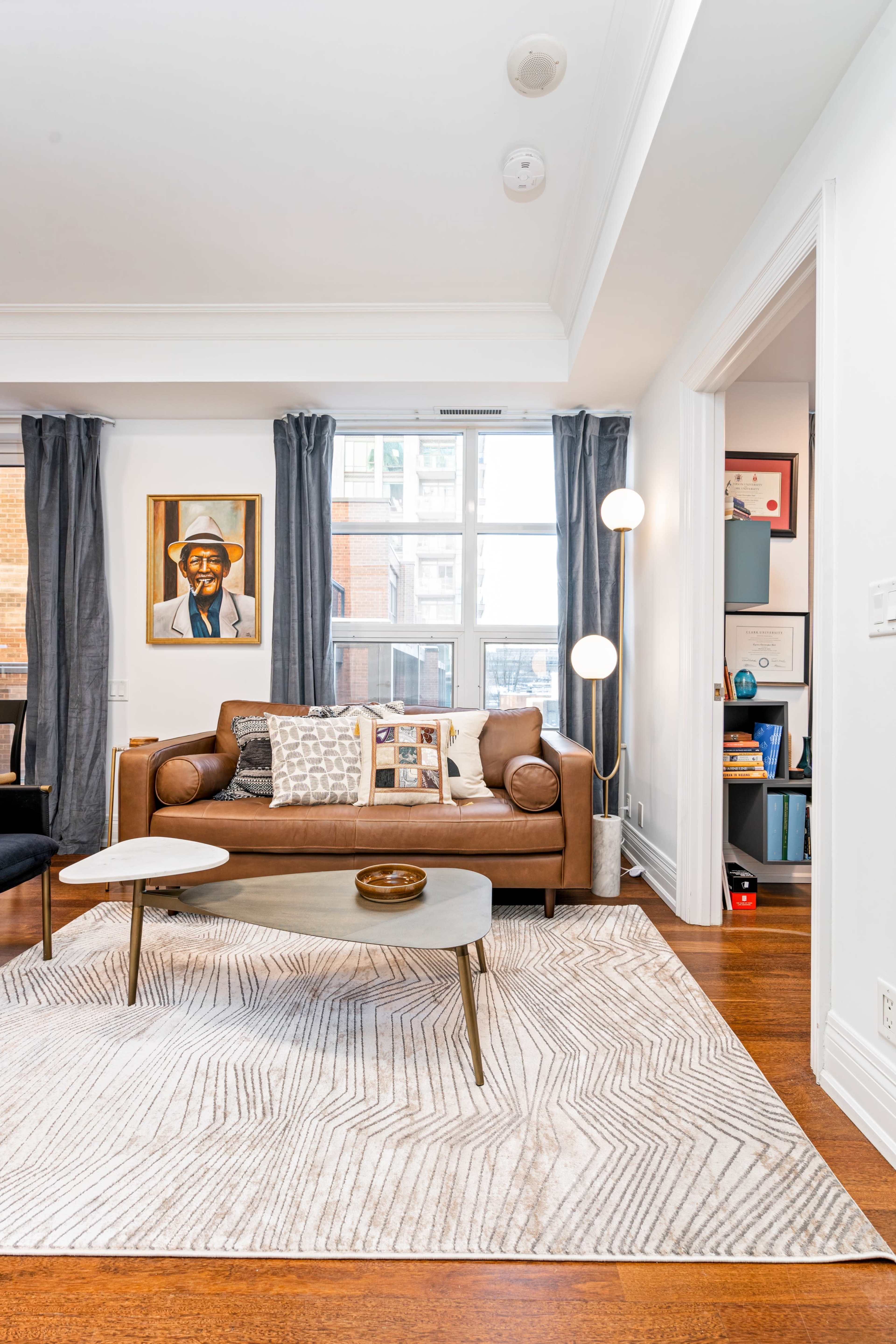 A modern living room featuring a brown leather sofa, a gray coffee table, and large windows with sheer curtains, all on a patterned rug.