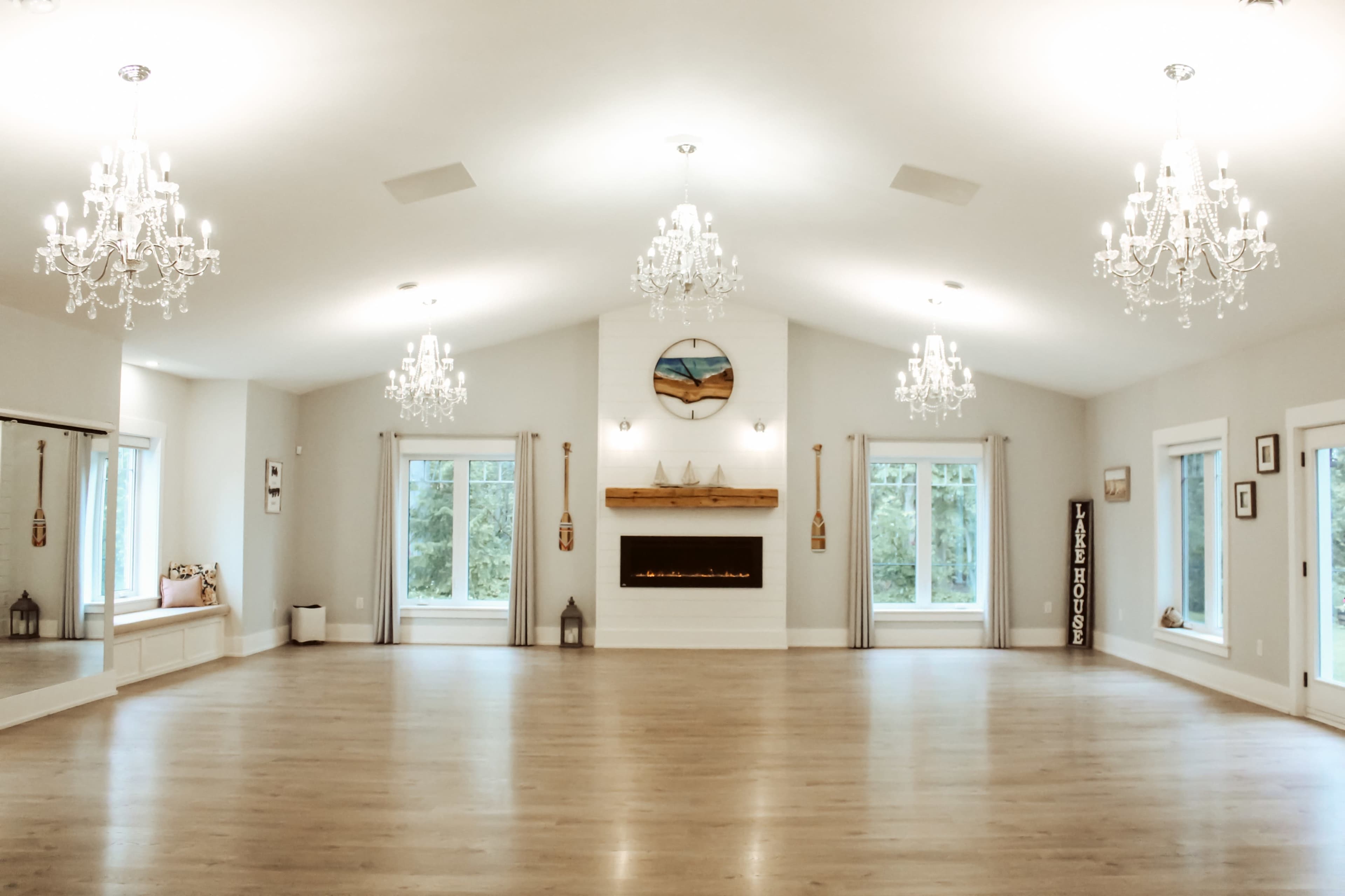 A spacious room with hardwood floors, large windows, a modern fireplace, and several chandeliers hanging from the ceiling.