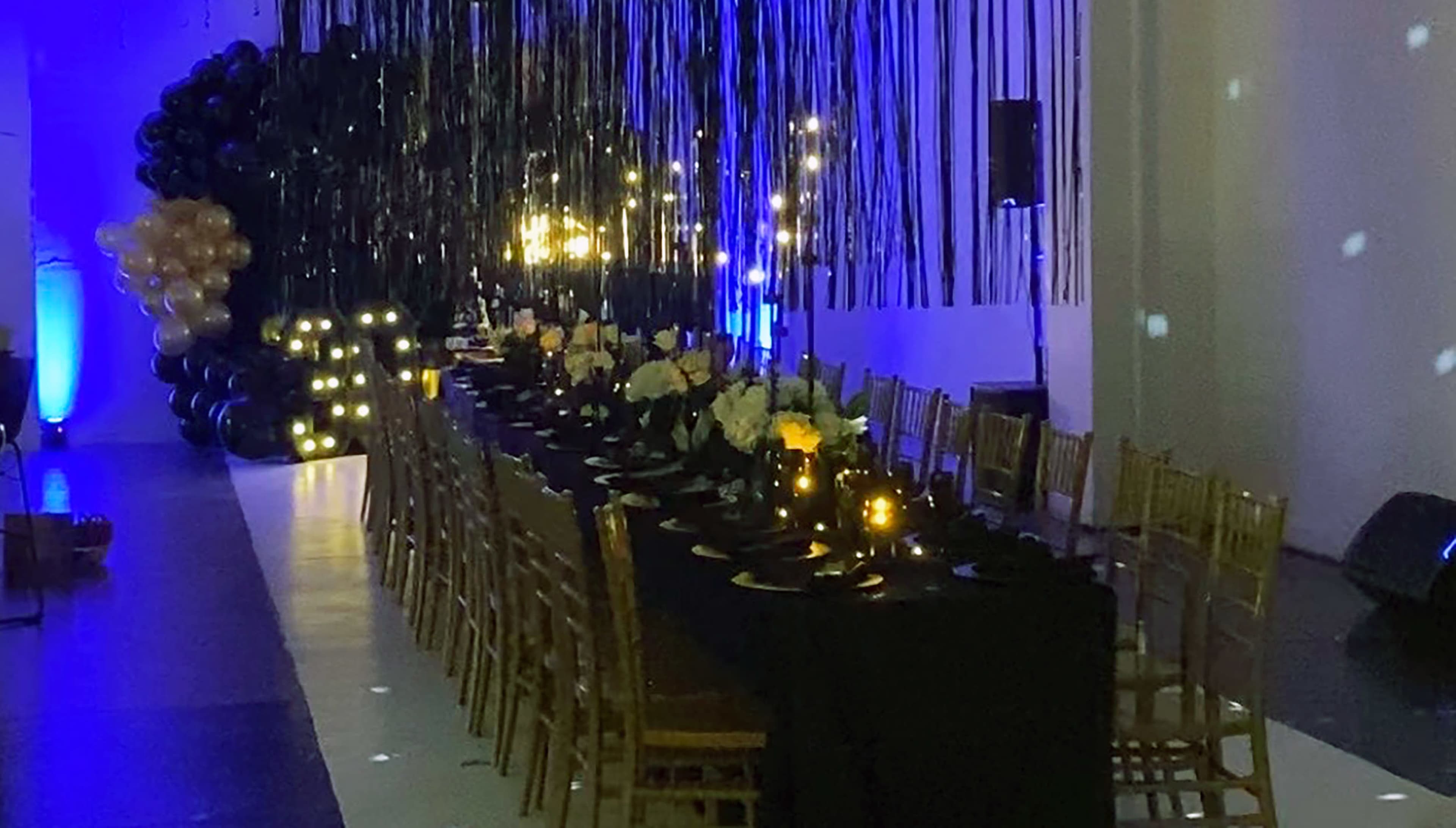 The image shows a long banquet table set with black tablecloths, lit by soft ambient lighting, and decorated with white flowers, surrounded by golden chairs in a dimly lit event space.