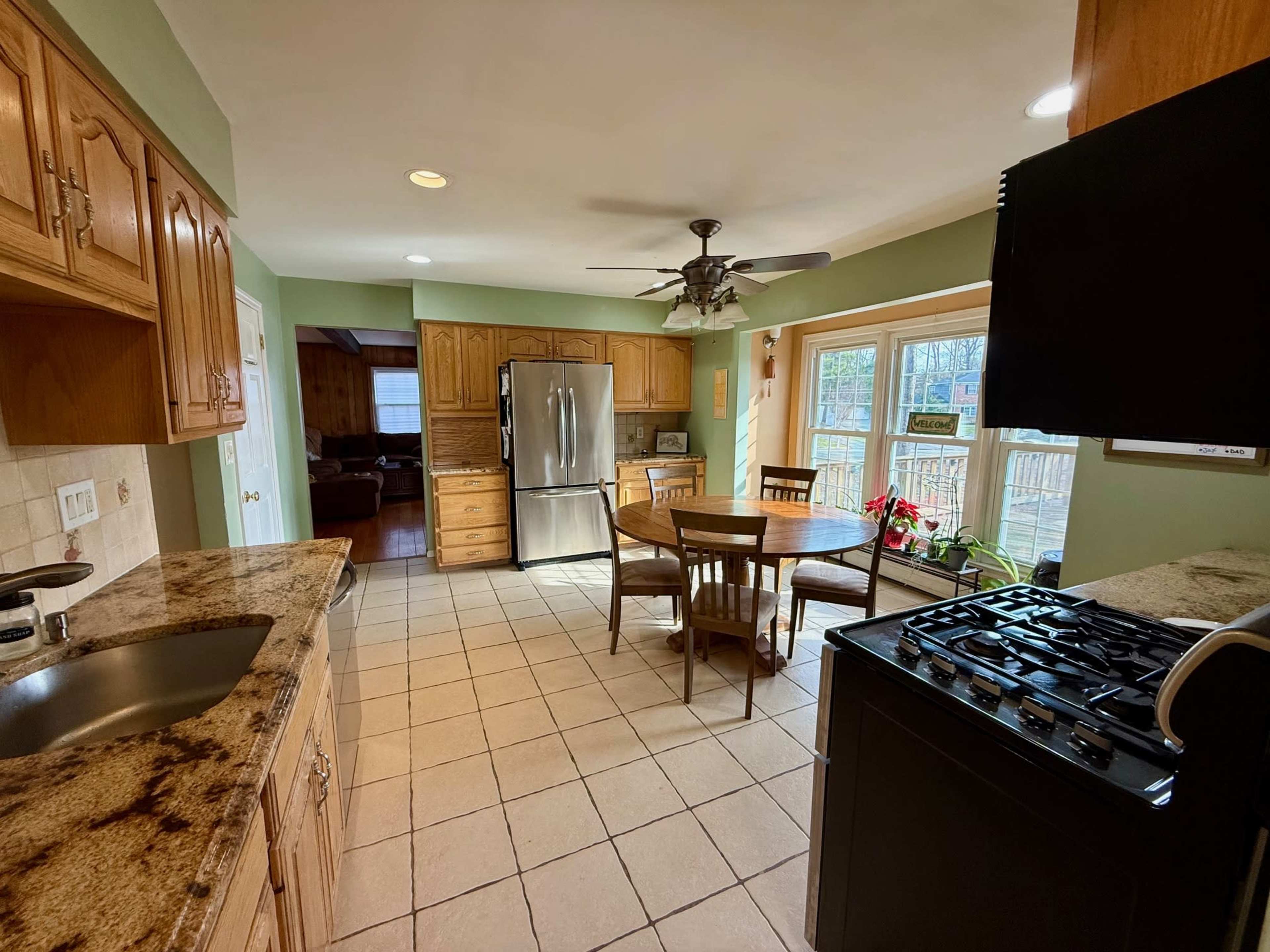 The image shows a kitchen with wooden cabinets, a stainless steel refrigerator, a dining table with chairs, and a view of a living room area through a doorway.