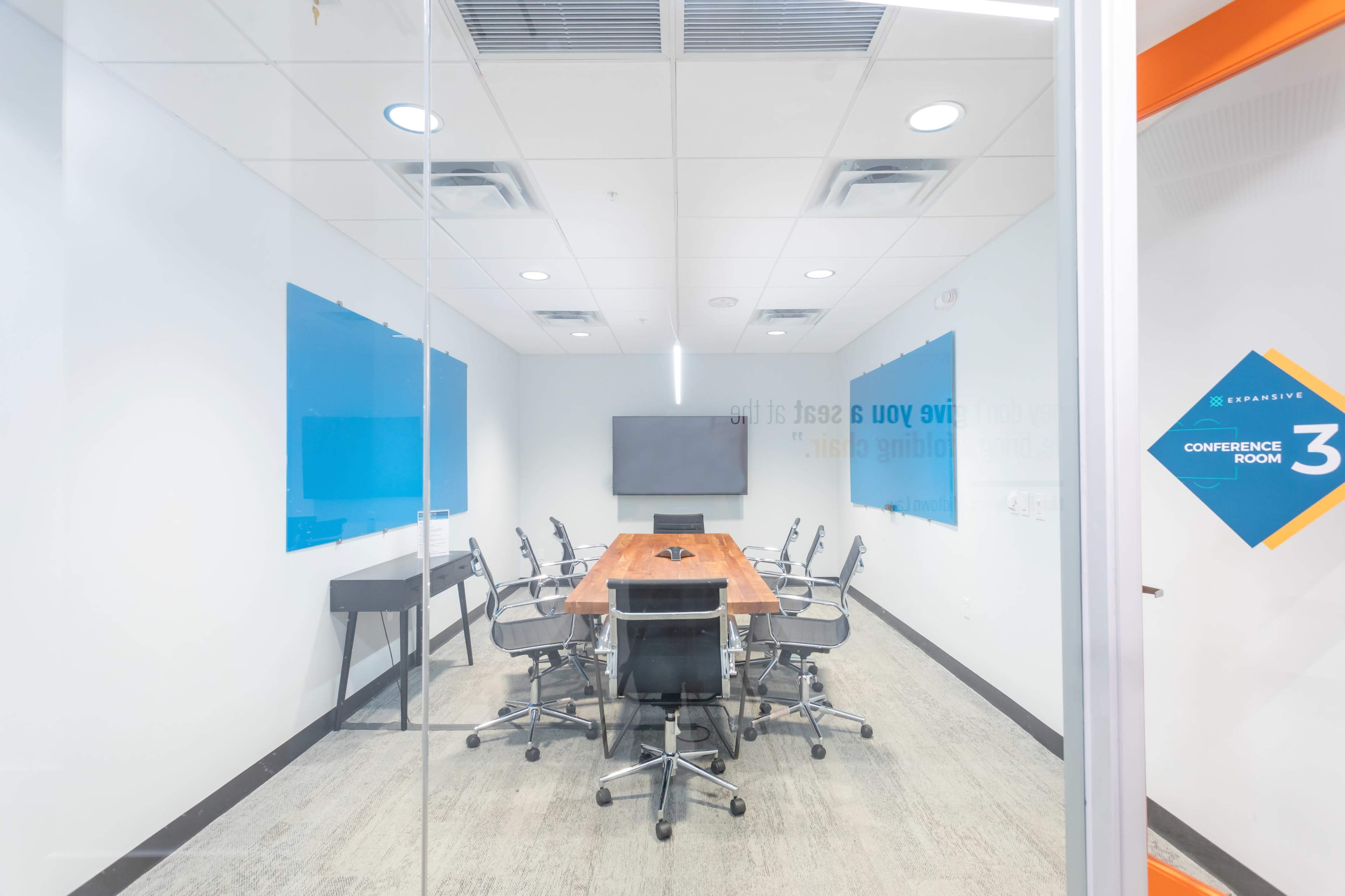 The image shows a modern conference room with a long wooden table, ergonomic chairs, a wall-mounted screen, and blue wall panels, all enclosed in glass.