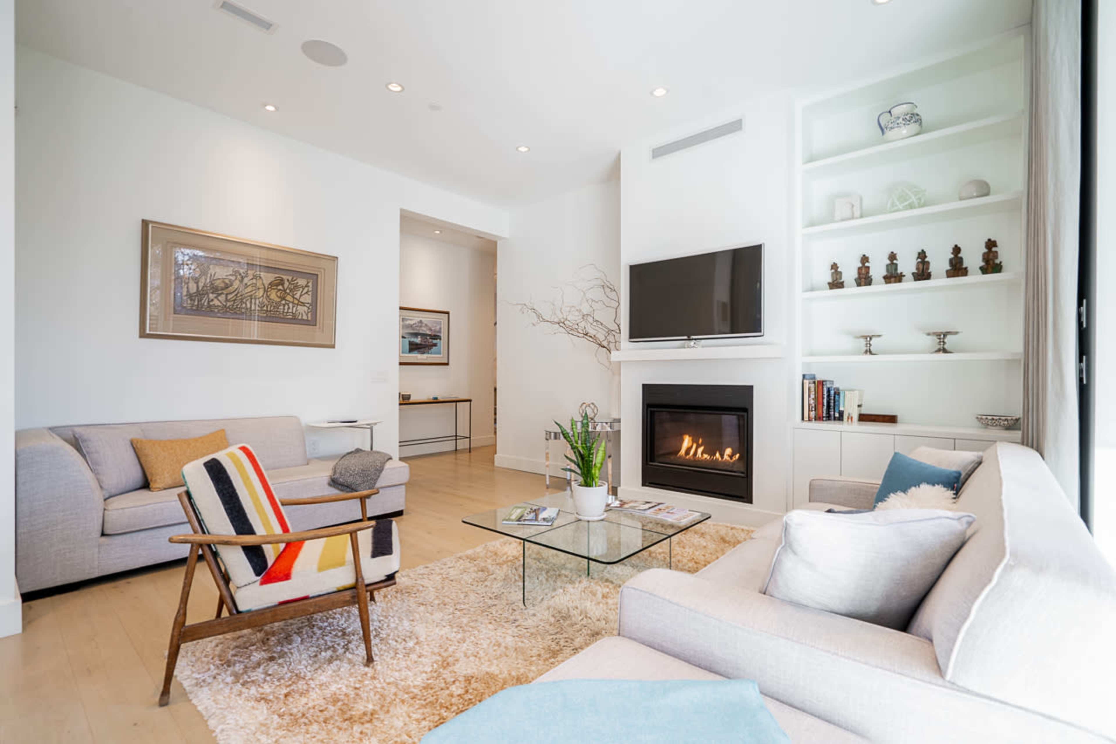The image shows a modern living room with a light color palette, a fireplace, a TV mounted on the wall, and seating arranged around a glass coffee table on a soft rug.