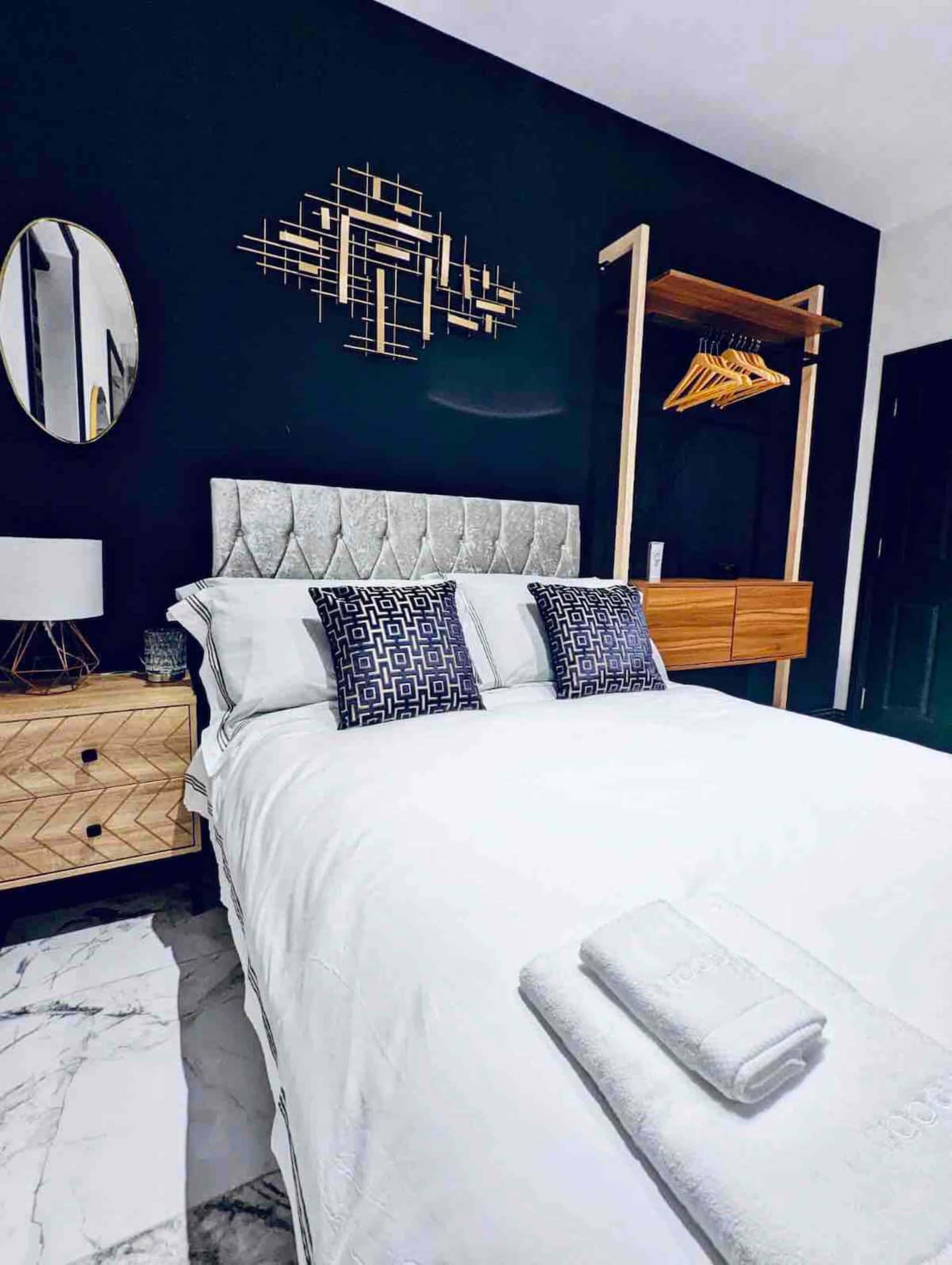 The image shows a modern bedroom featuring a bed with decorative pillows, a wooden nightstand, a hanging wardrobe, and dark walls.