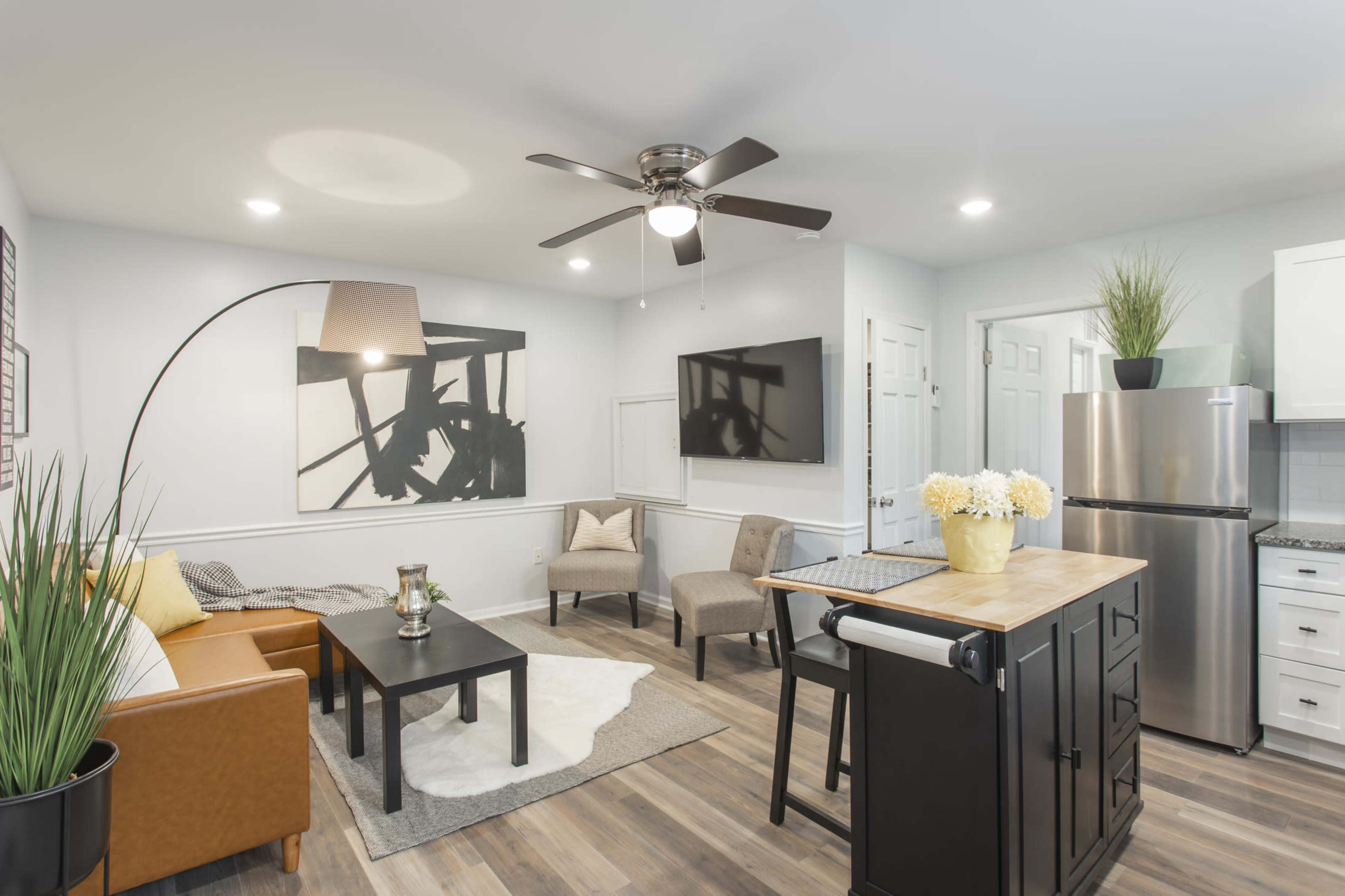 A modern living space featuring a brown leather couch, a black coffee table, a kitchen island, and a ceiling fan, with a mounted flat-screen TV on the wall.