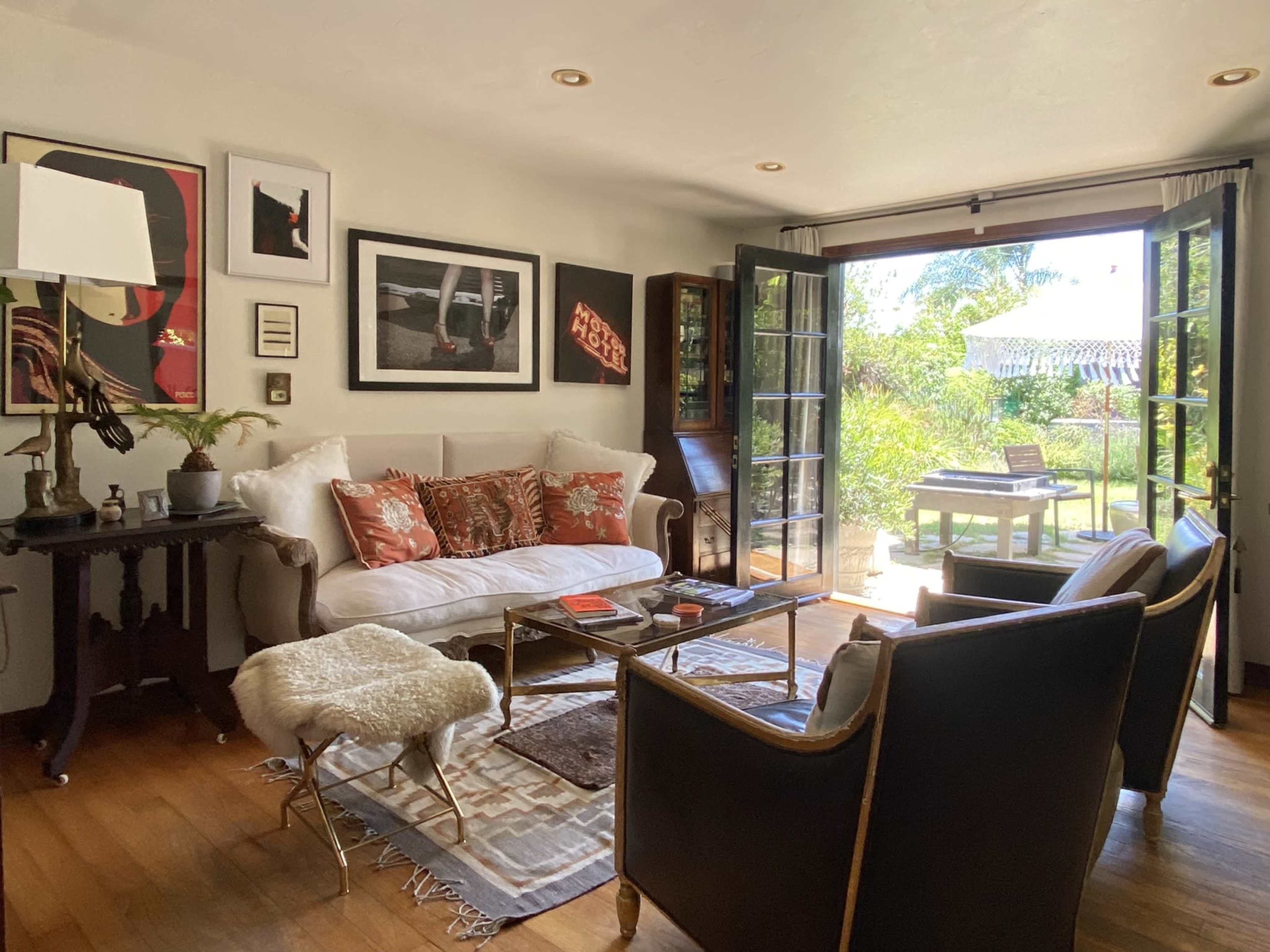 The image shows a living room with a white sofa, two dark armchairs, a glass coffee table, and large doors leading to a garden area.