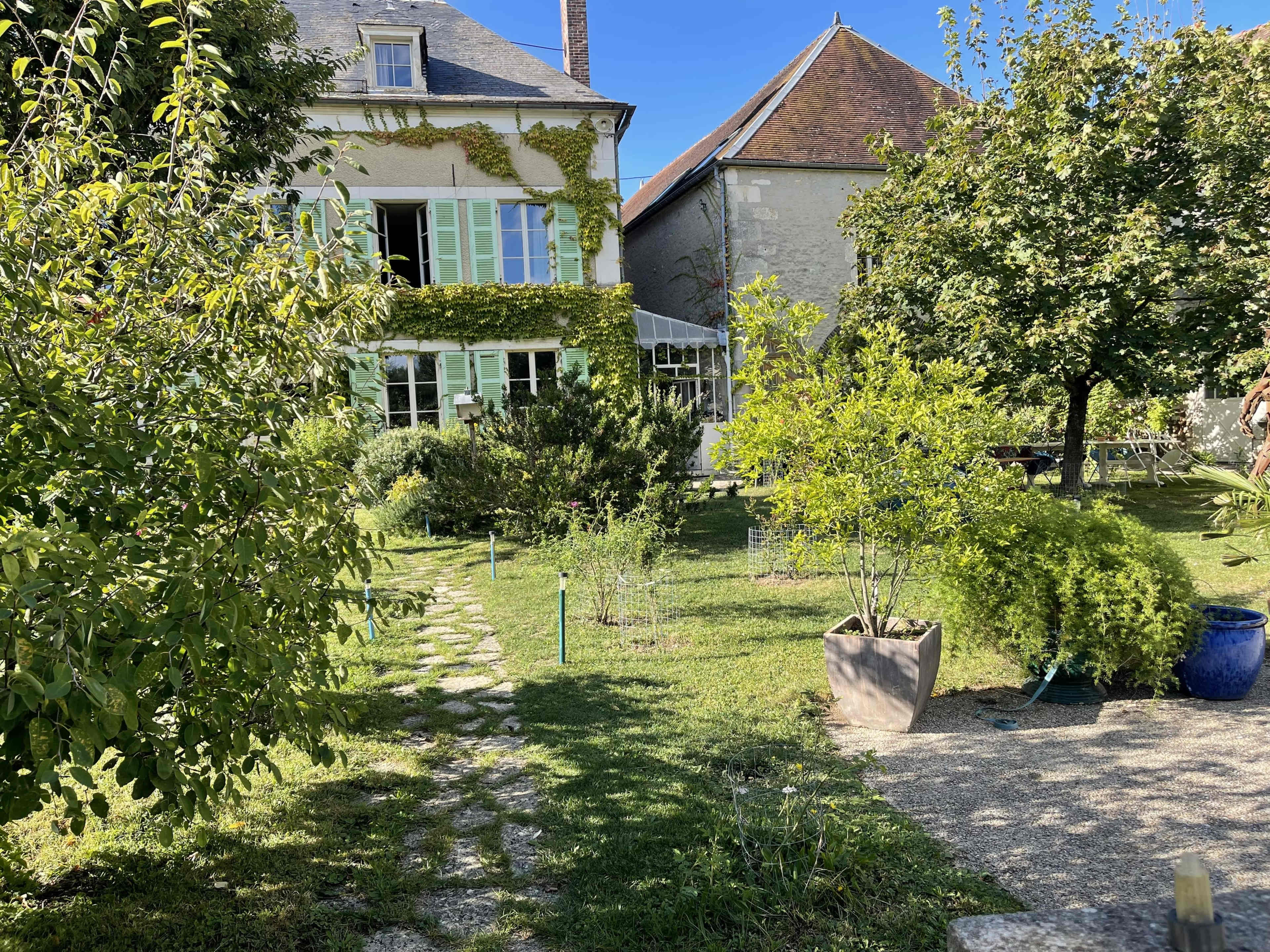 The image shows a green garden with walking paths, featuring a house adorned with vines, surrounded by various plants and trees under a clear blue sky.