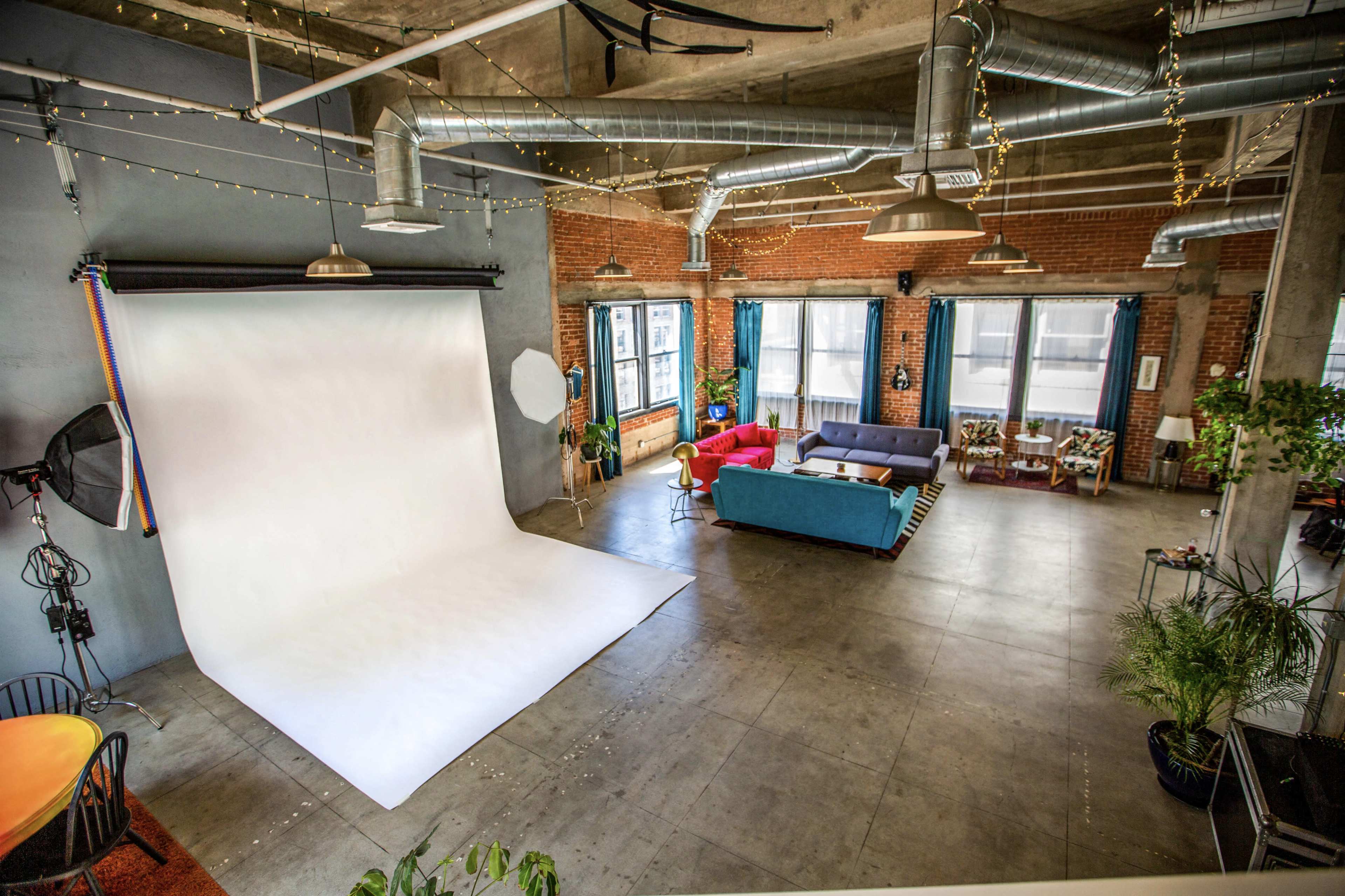 A spacious loft with a photography backdrop, seating area, and various plants, illuminated by large windows.