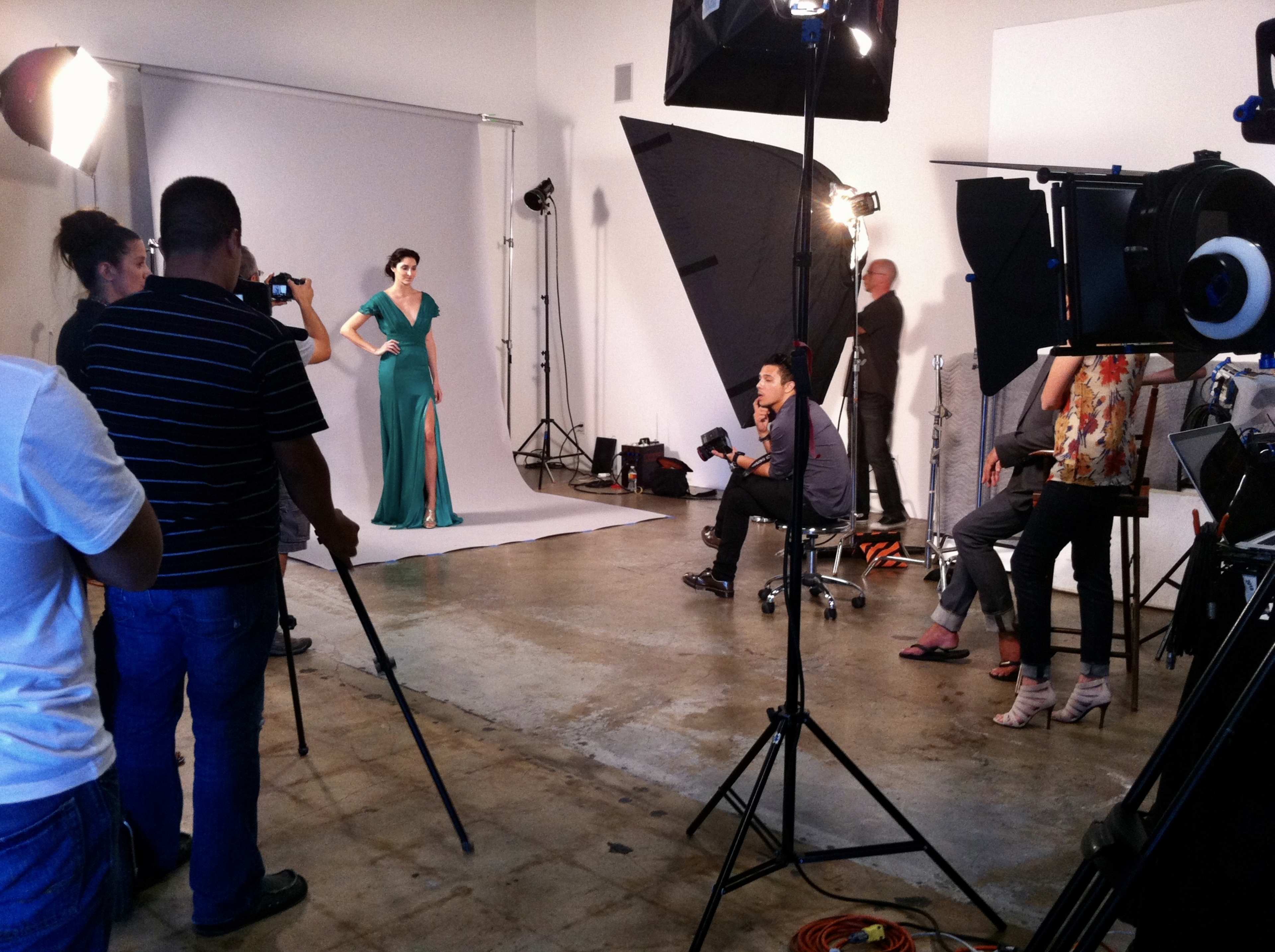 A model poses in a green gown while photographers and crew members work in a studio setting.