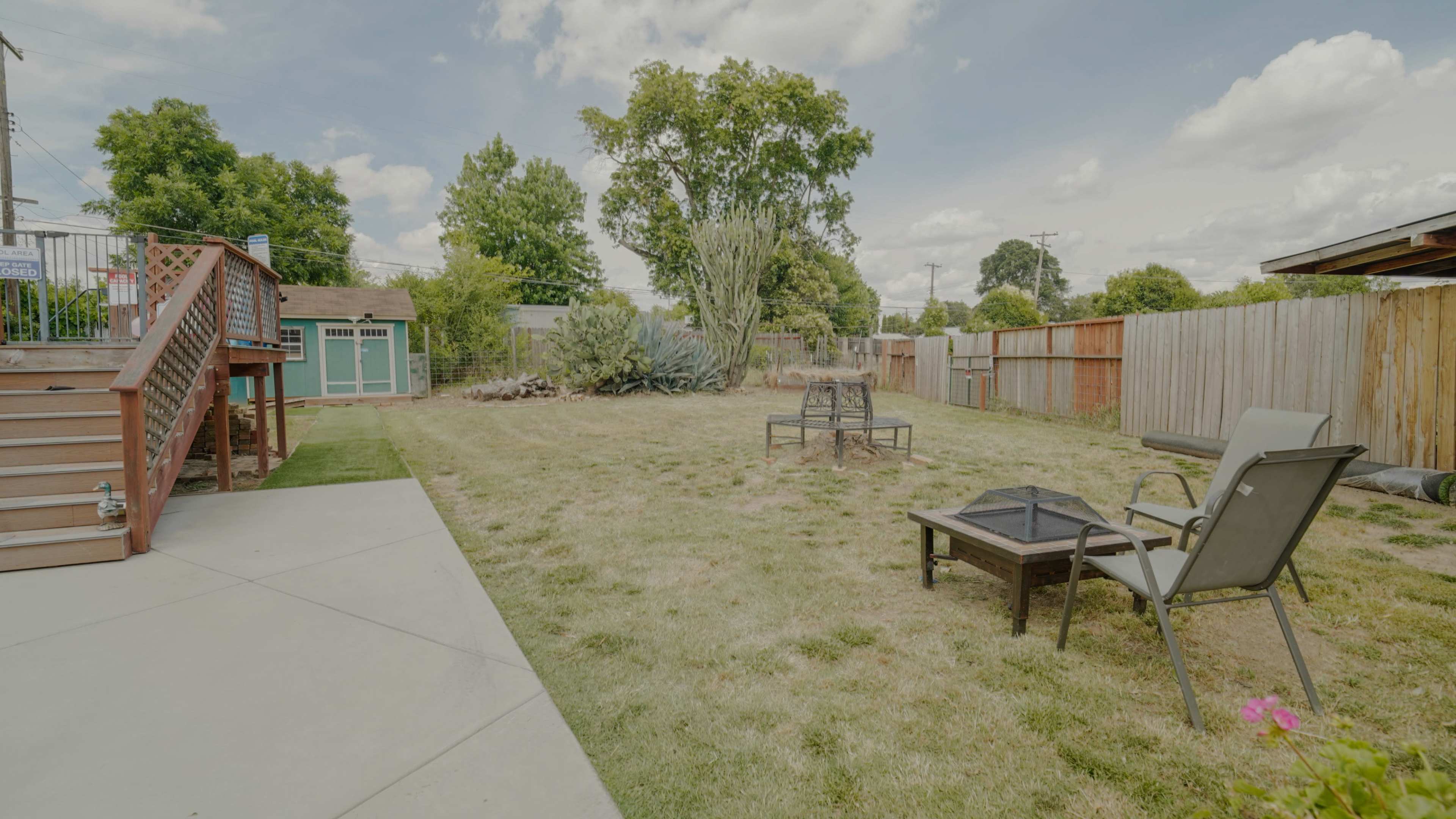 The image shows a spacious backyard with a fire pit, a seating area, and greenery surrounded by wooden fences.