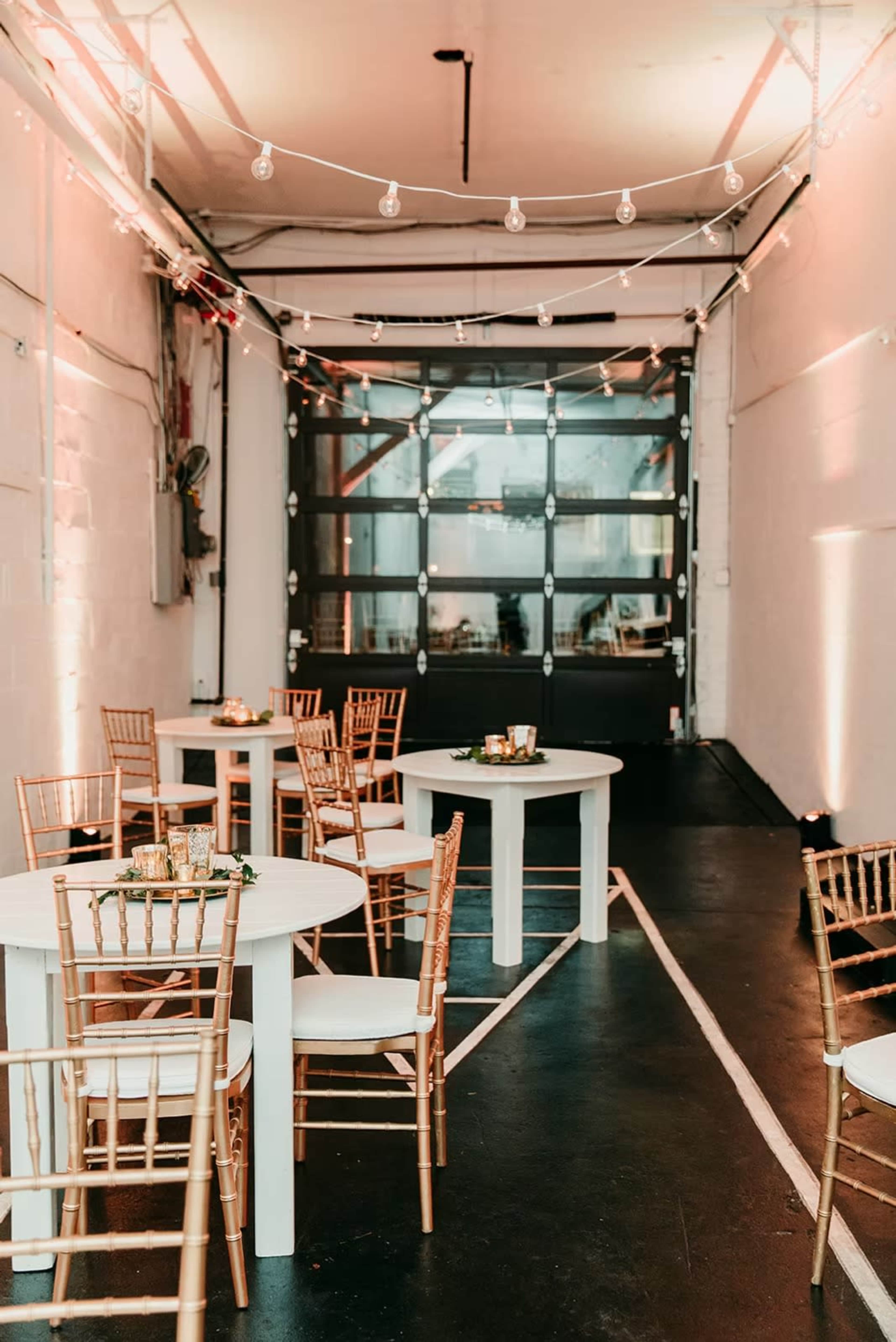 The image shows an indoor event space featuring round tables with gold chairs, string lights overhead, and a large door at the far end.