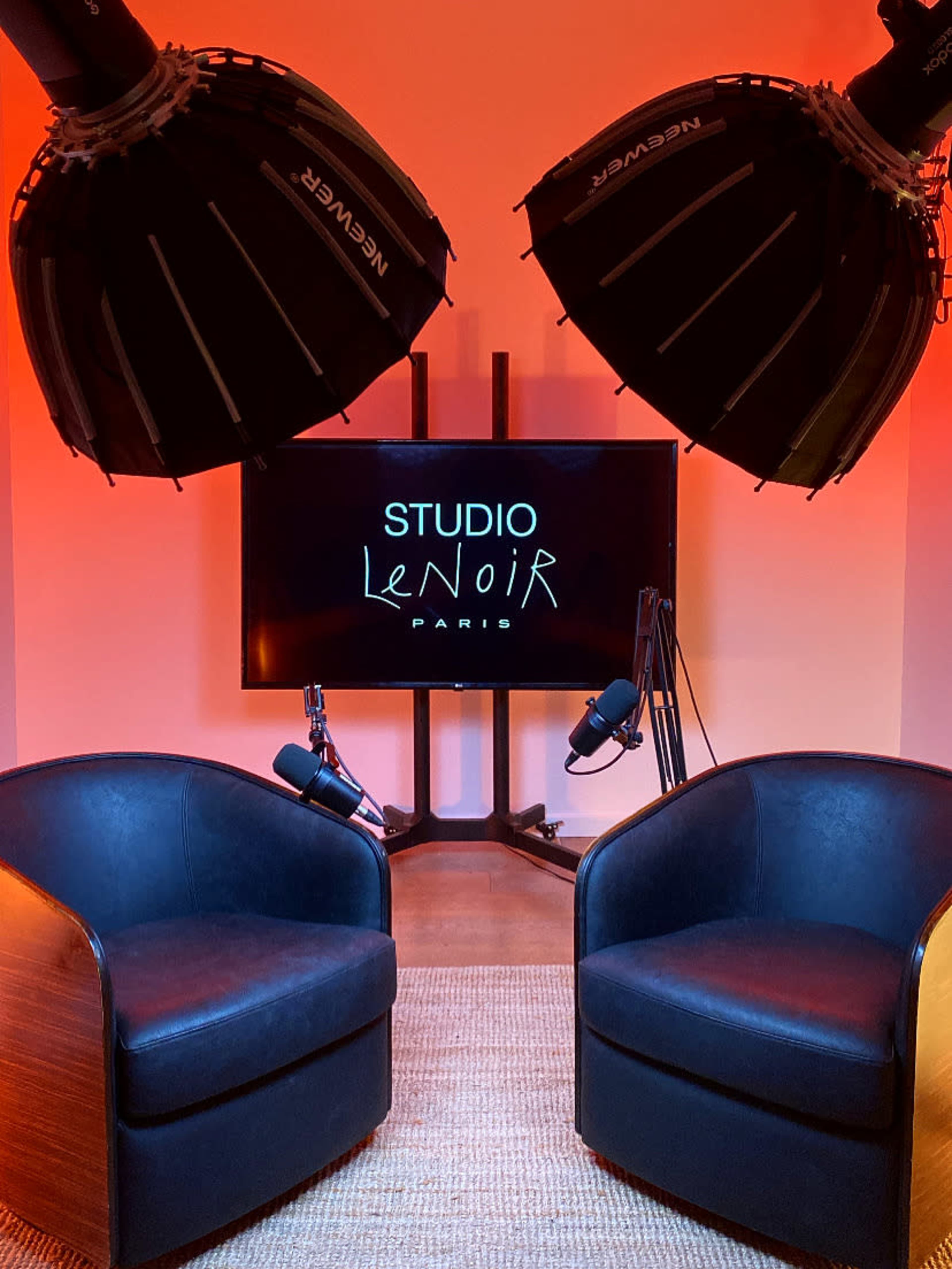 The image shows a studio setting with two black leather armchairs facing a large screen displaying the words "STUDIO Le NOIR PARIS," illuminated by two large softbox lights overhead.