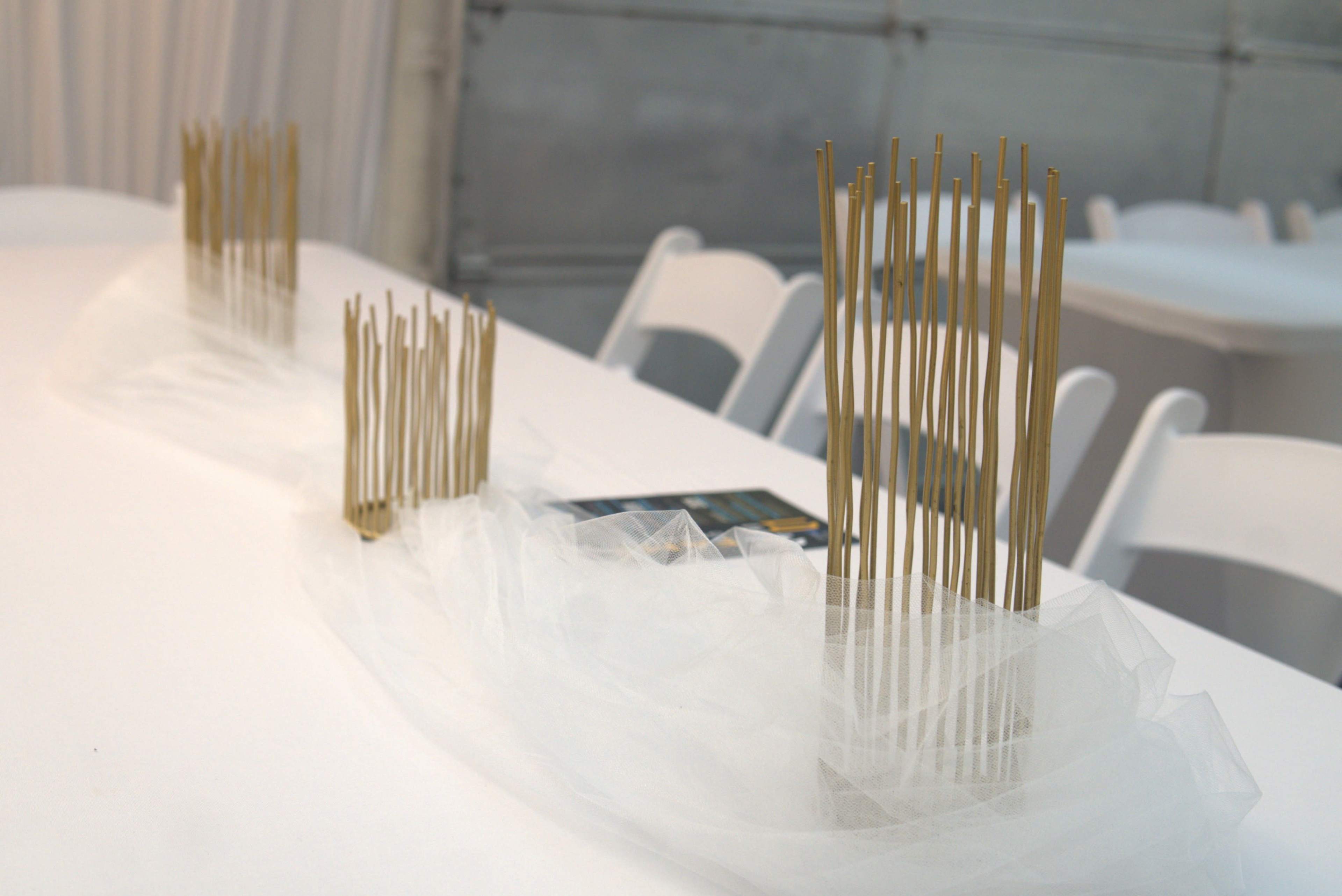 Two arrangements of golden sticks are placed on a table draped with white fabric in a tented setting.
