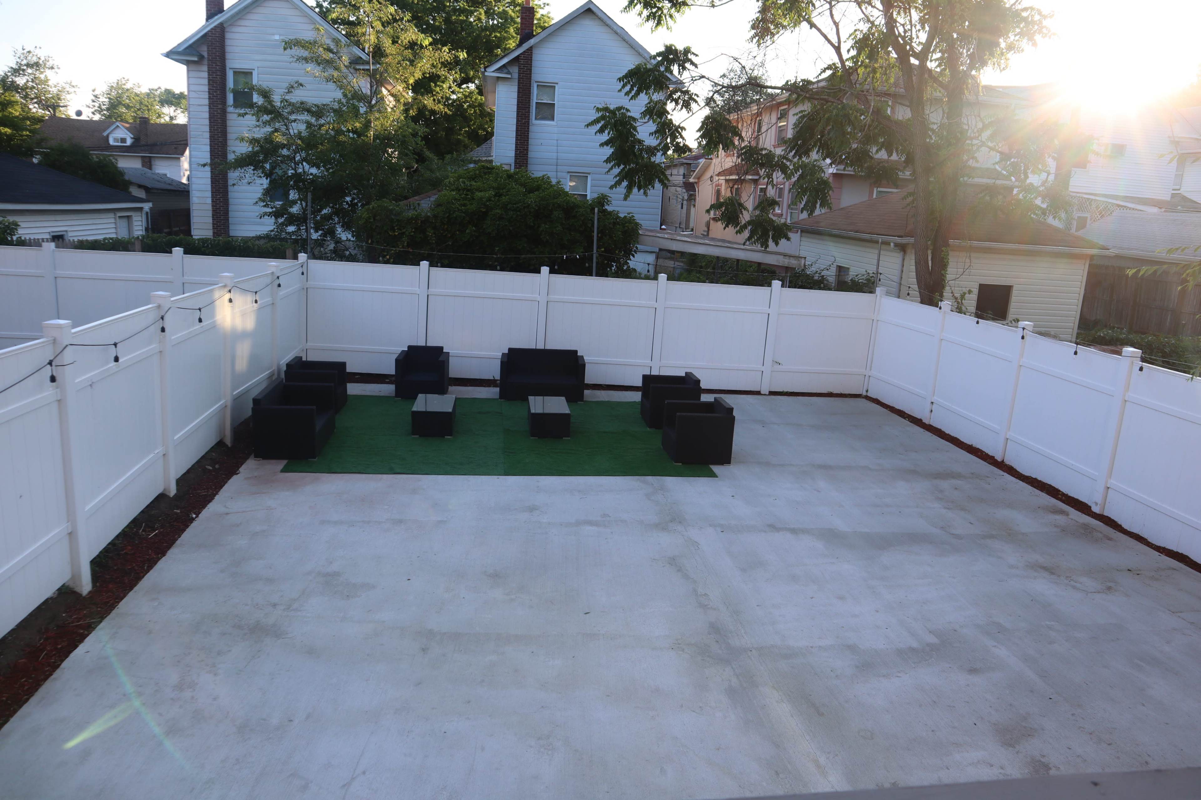 A concrete backyard area enclosed by a white fence, featuring a green patch of artificial grass and several black outdoor seating arrangements.