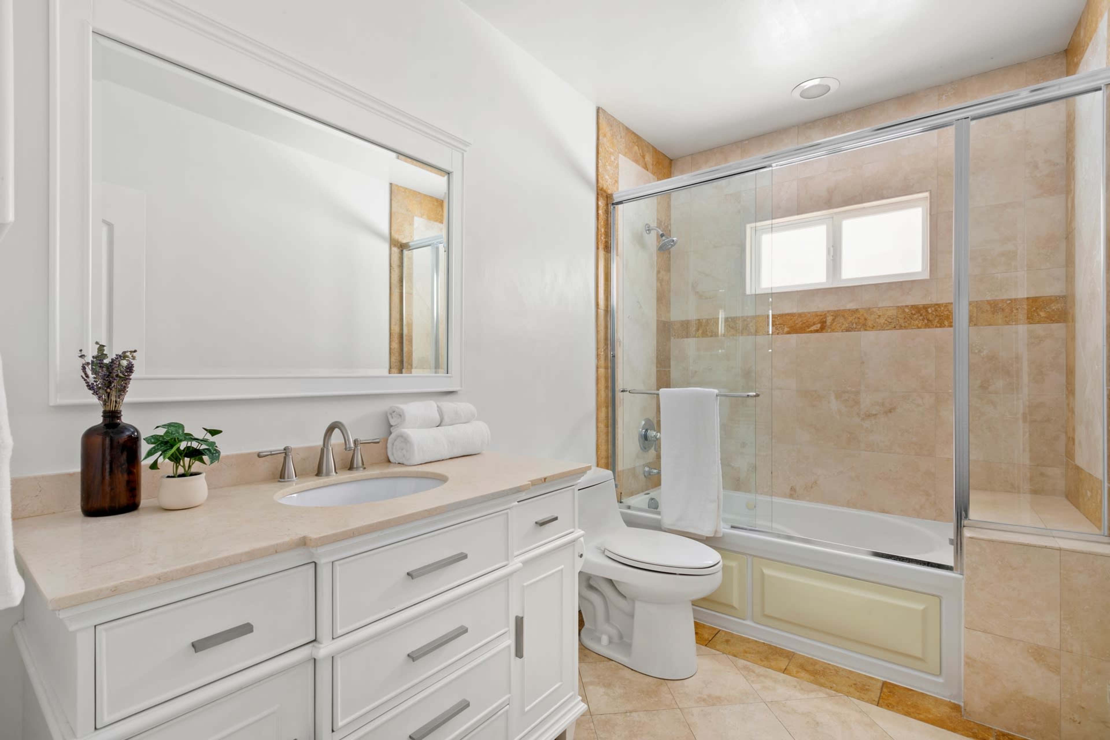 The image shows a modern bathroom featuring a glass-enclosed shower, a white vanity with a sink, a toilet, and a large mirror.