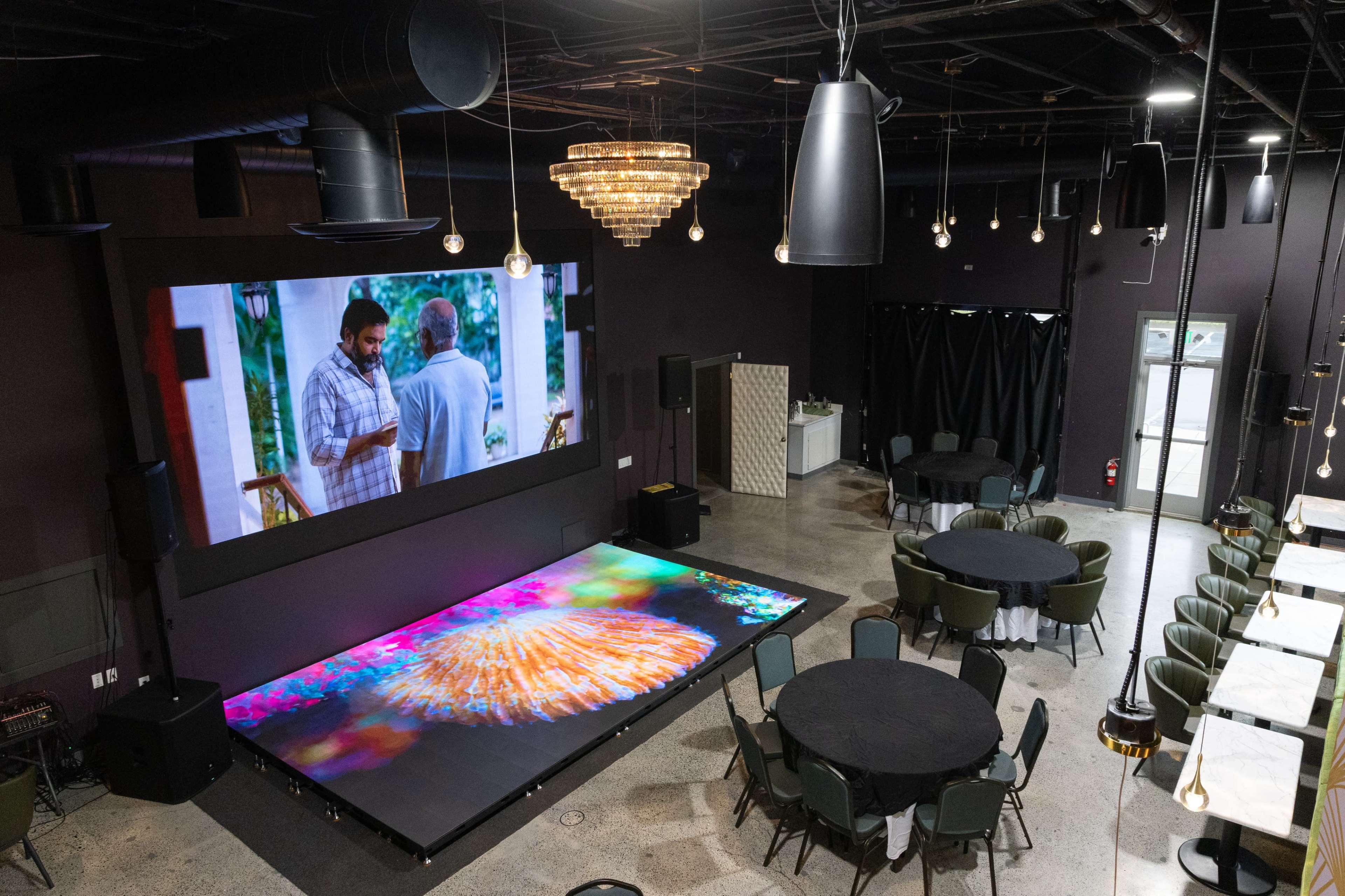 The image shows an indoor event space with a large screen displaying a video, round tables set up in the foreground, and modern lighting fixtures hanging from the ceiling.