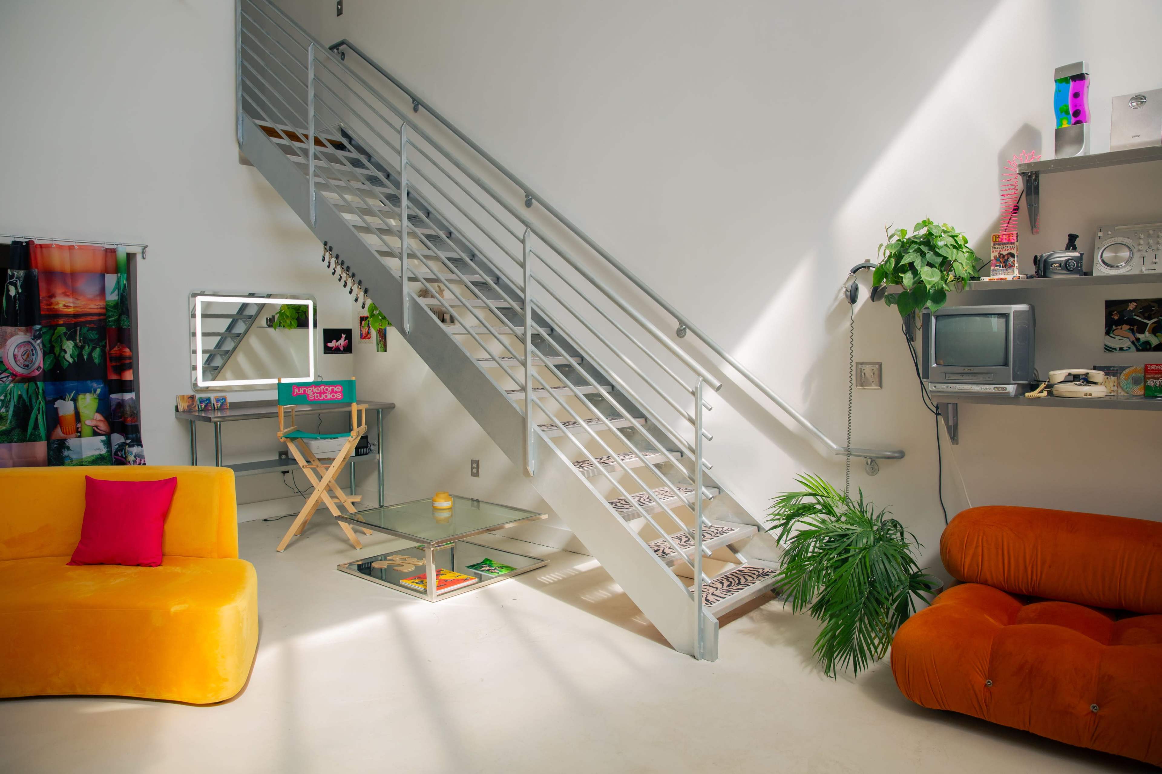 The image shows a modern living space with a staircase, a glass coffee table, a mirror, and colorful furniture, including a bright orange chair and a pink cushion.