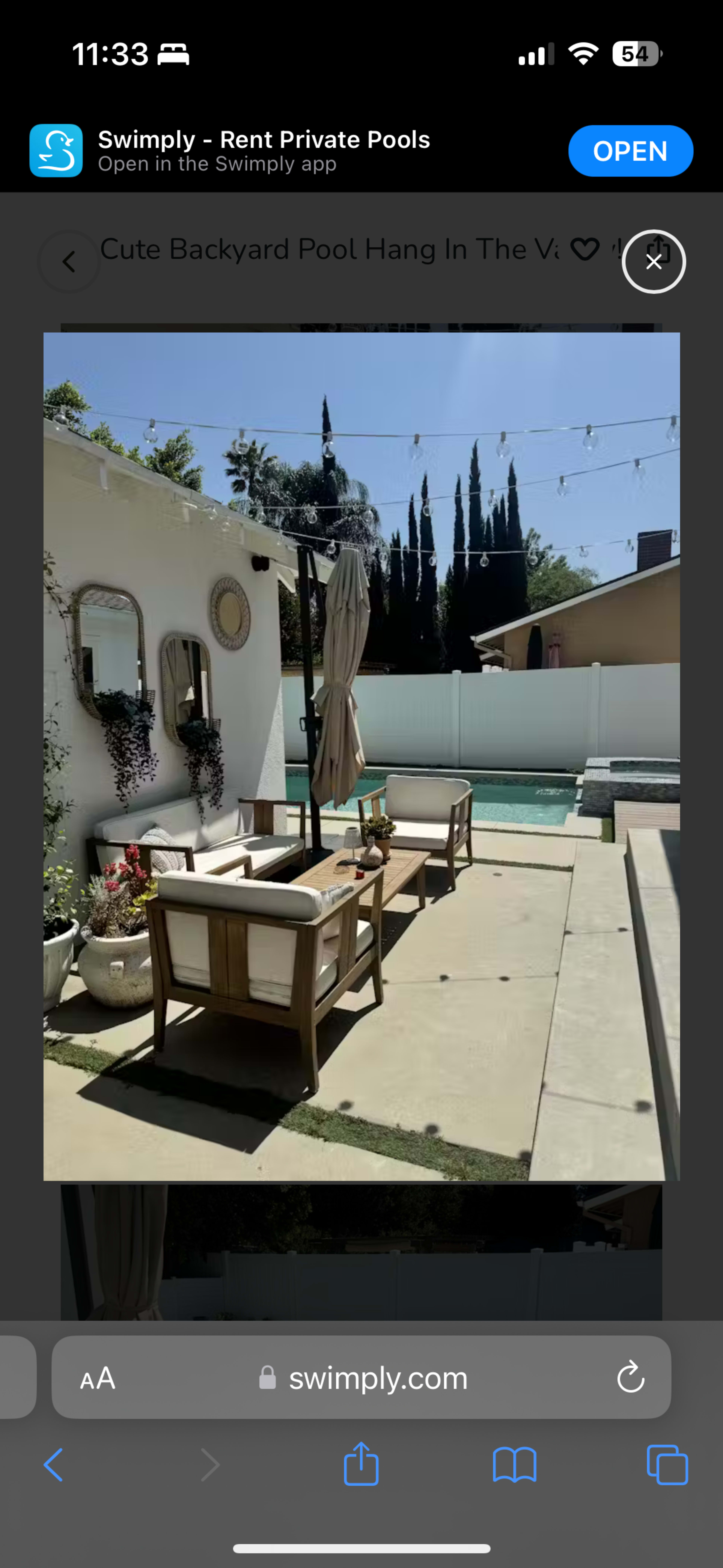 A backyard pool area features a seating arrangement with two chairs and a table under a parasol, surrounded by greenery and a swimming pool.