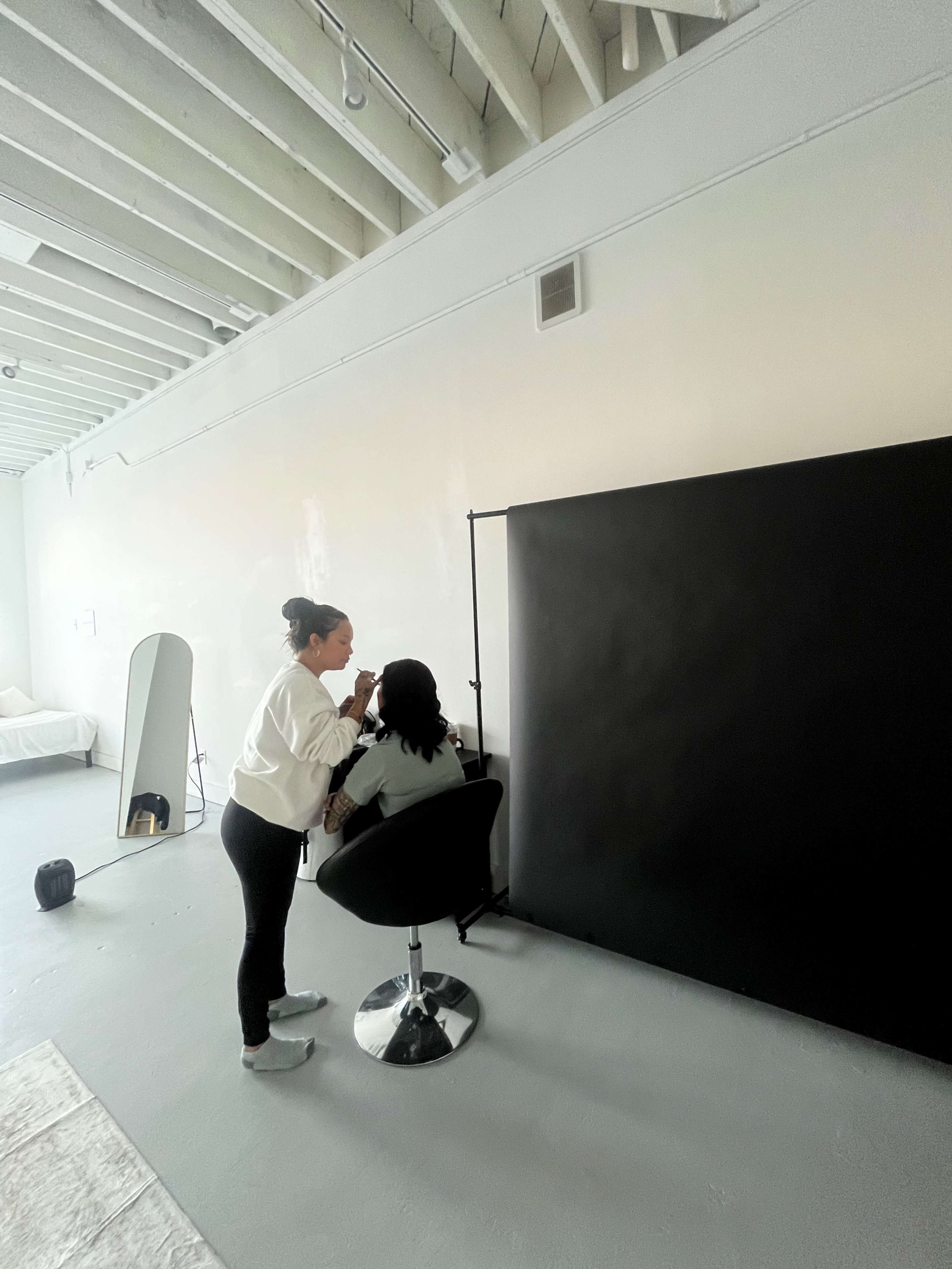 A person is applying makeup to another individual who is seated in a salon chair in a minimalist, brightly lit studio space.
