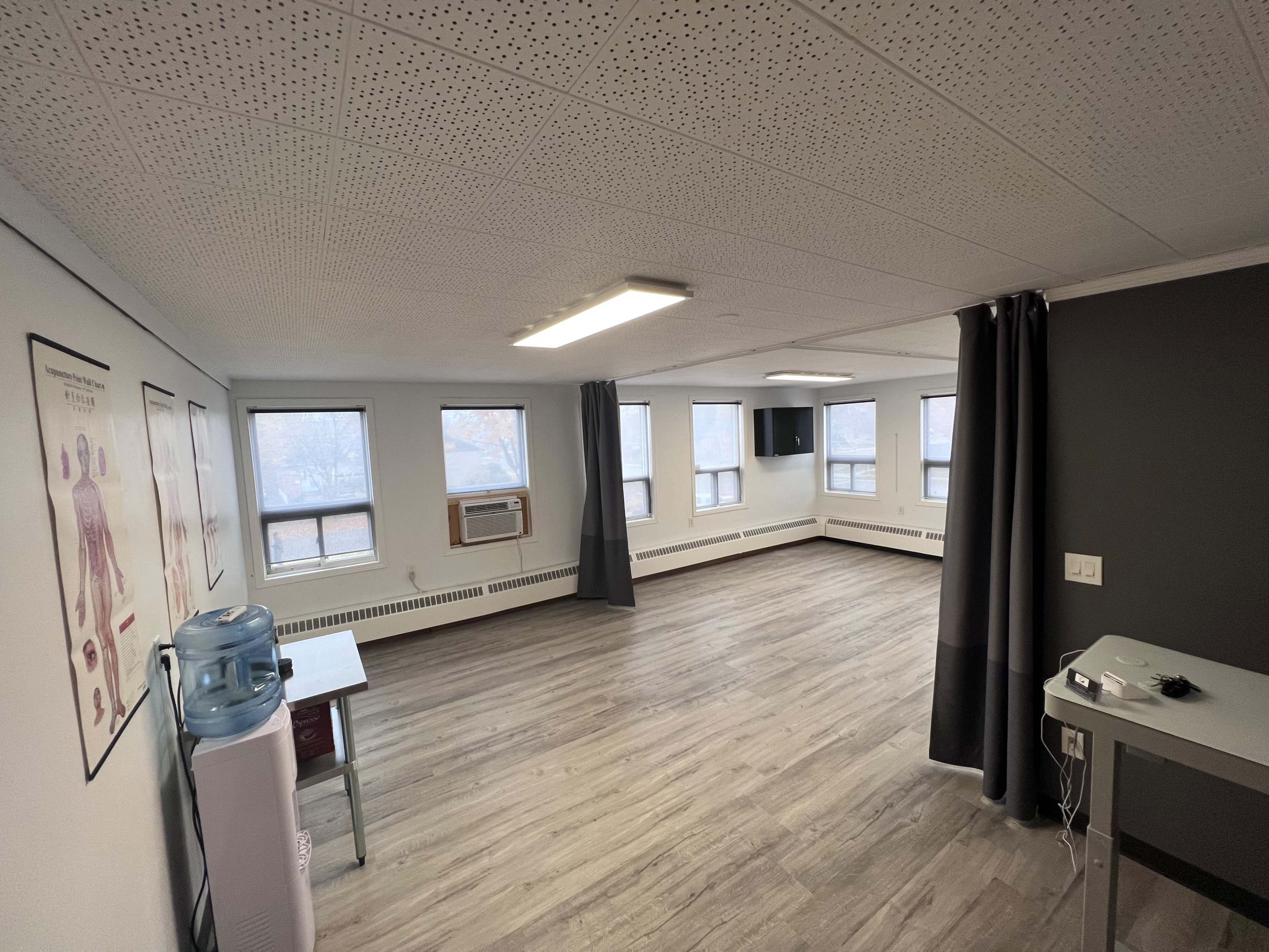 An empty room with wooden flooring, multiple windows, a wall-mounted television, and medical posters hanging on the walls.