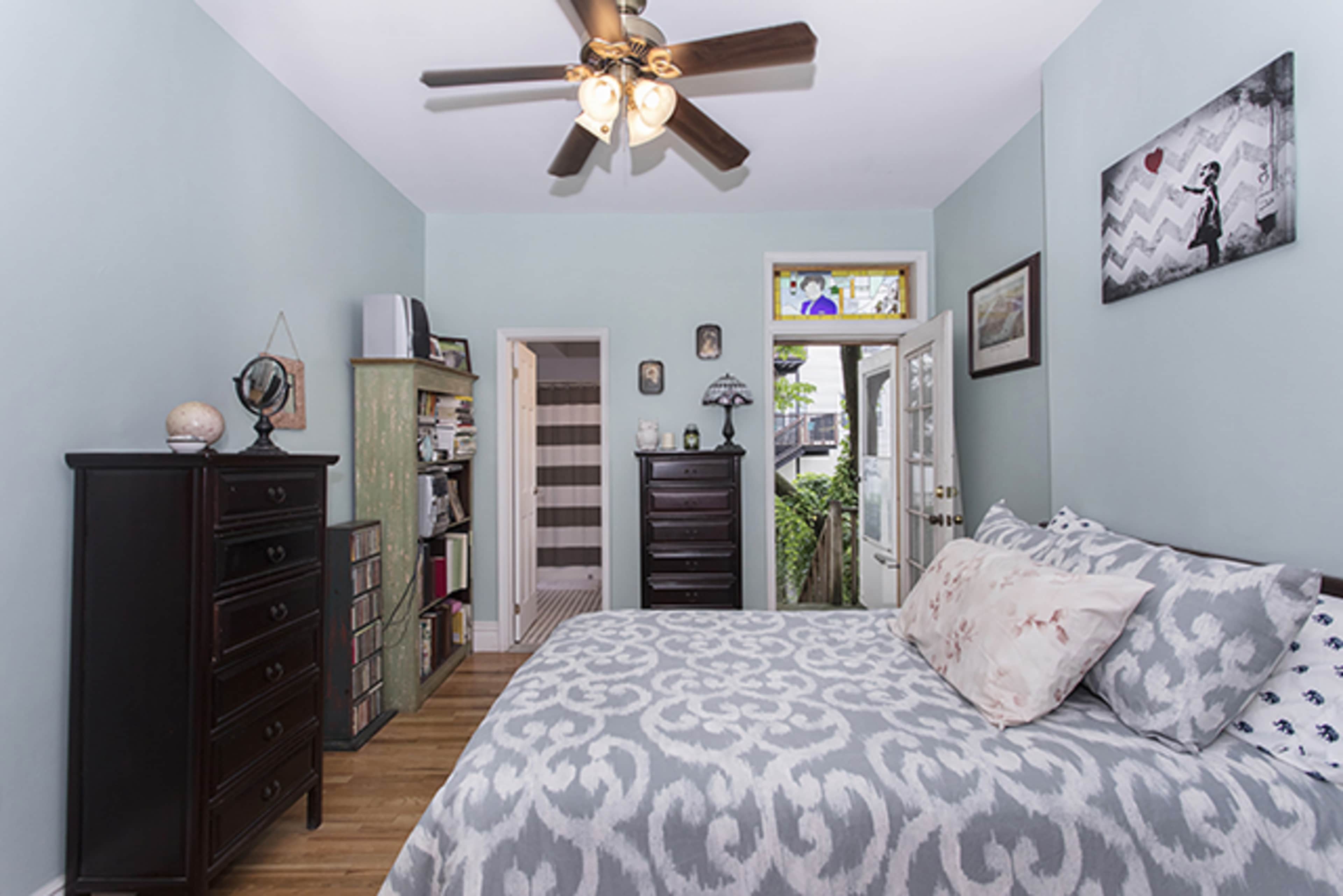 The image shows a bedroom with a bed, a dresser, and a ceiling fan, featuring light blue walls and a door leading to an outdoor area.