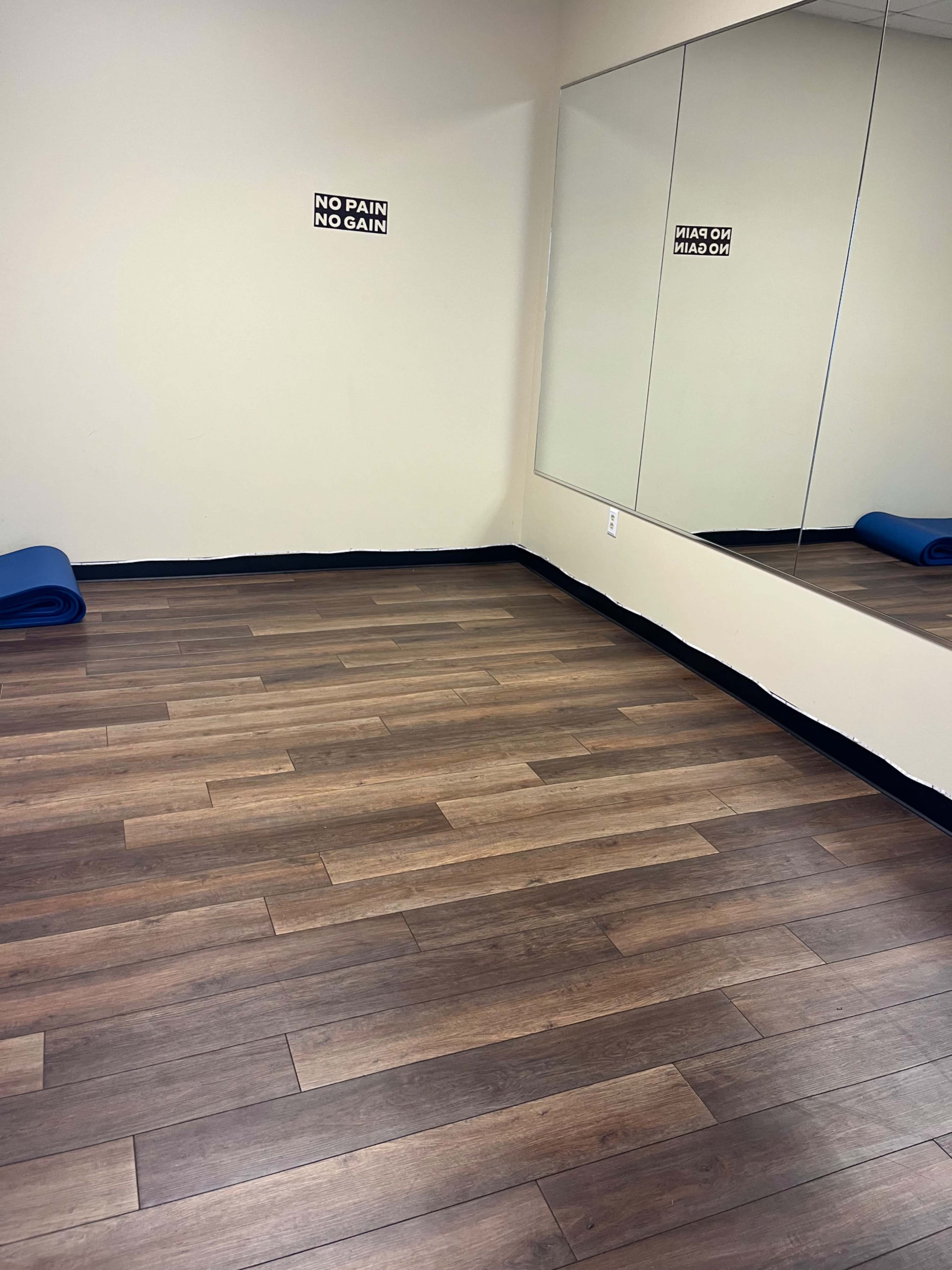 The image shows a fitness room with wooden flooring, two yoga mats rolled up against the wall, and motivational signs on the walls.