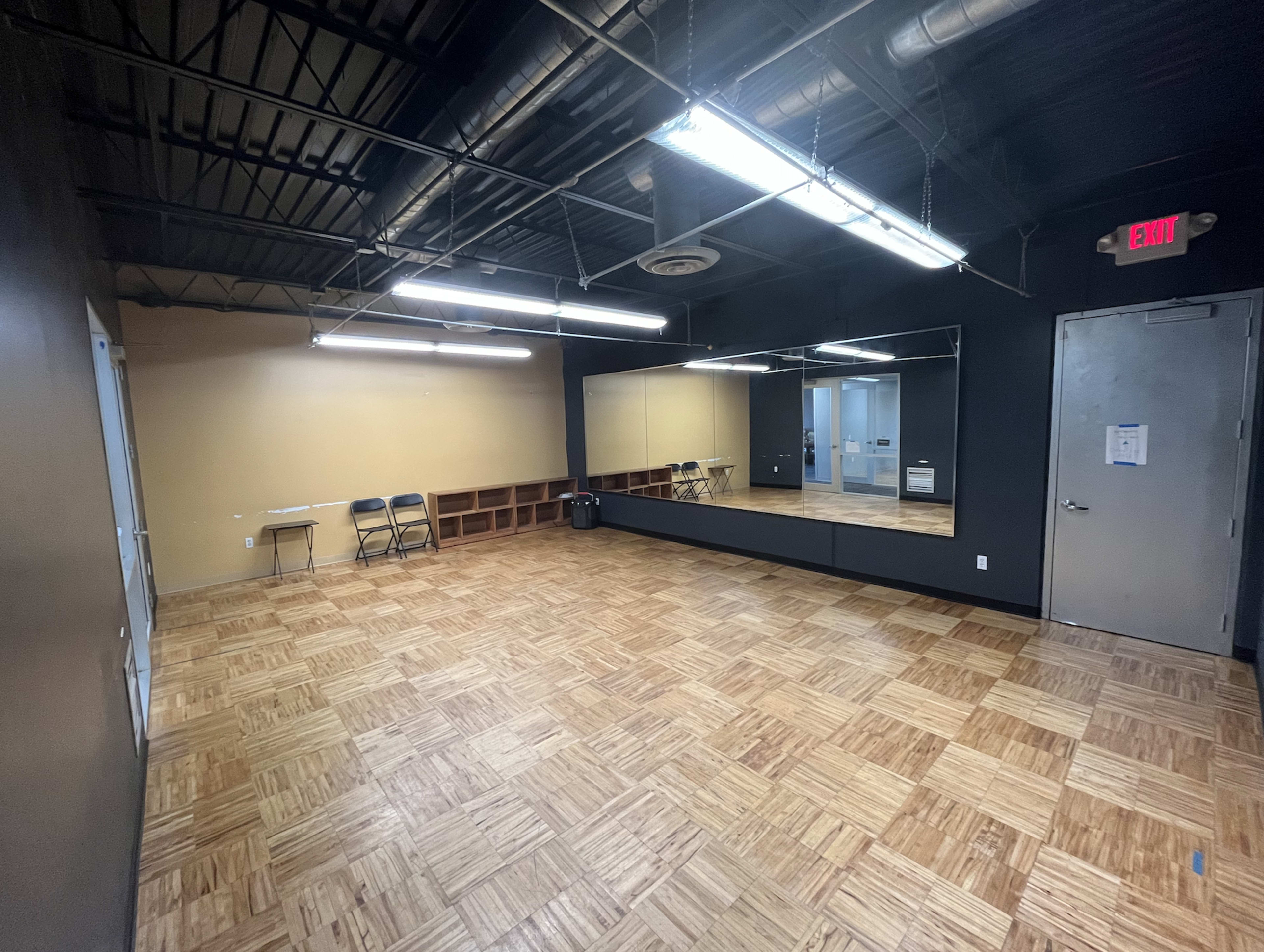 A spacious, empty room with a hardwood floor, black walls, overhead fluorescent lights, and a large mirror along one side.