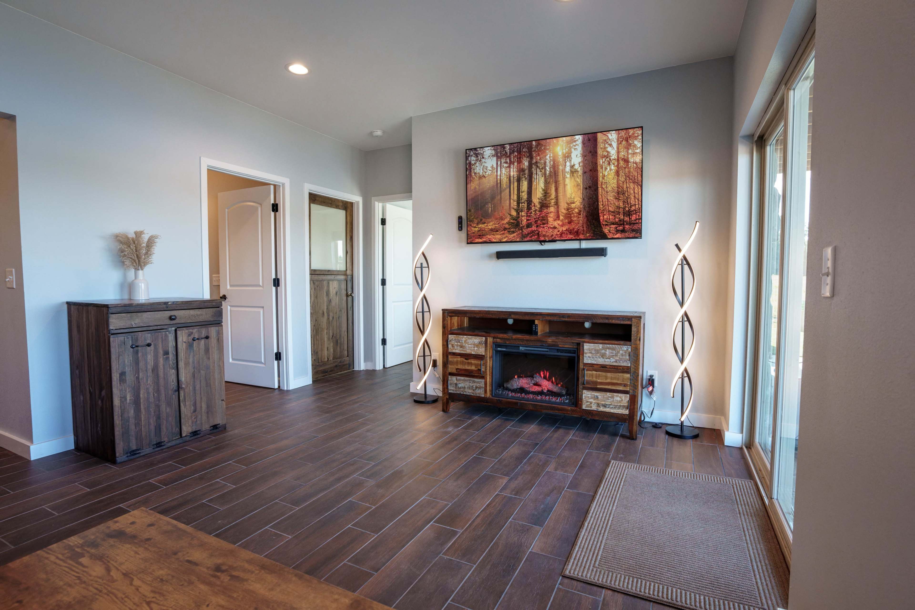 The image shows a modern living room with wood flooring, a mounted television displaying a forest scene, and contemporary décor, including floor lamps and a wooden console.