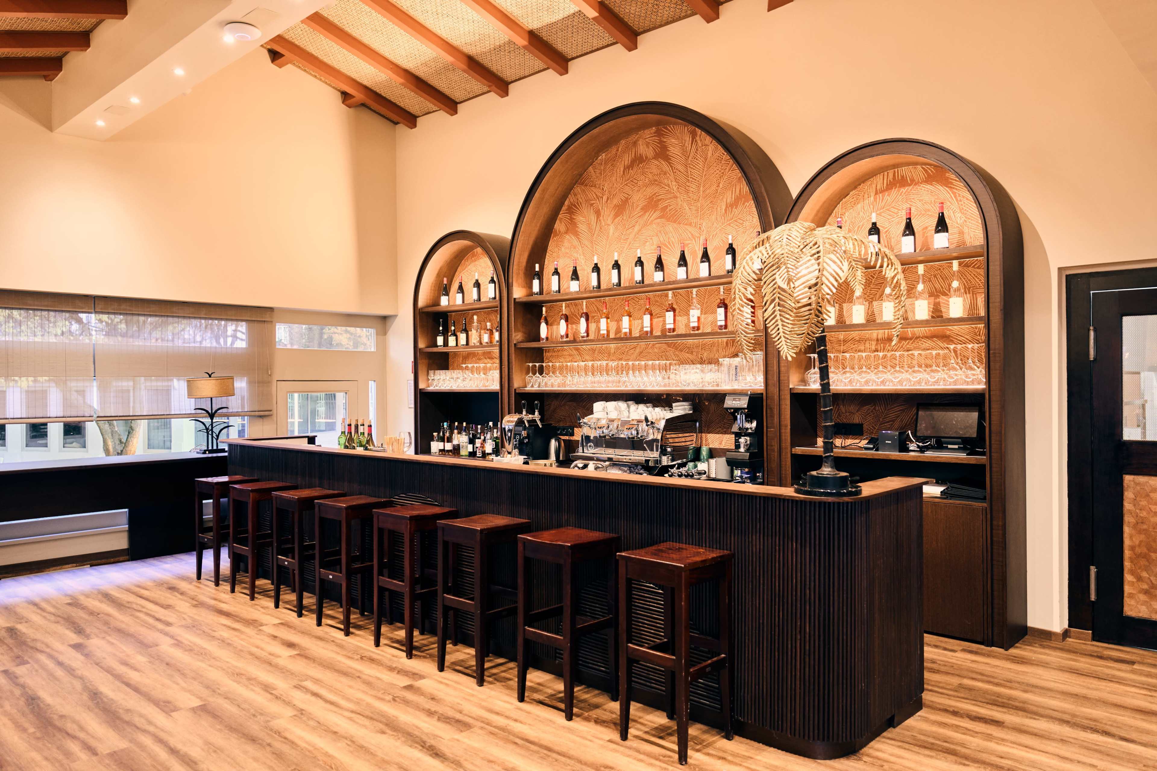 The image shows a modern bar with a wooden counter, several high chairs, and shelves displaying various bottles and glassware against a neutral backdrop.
