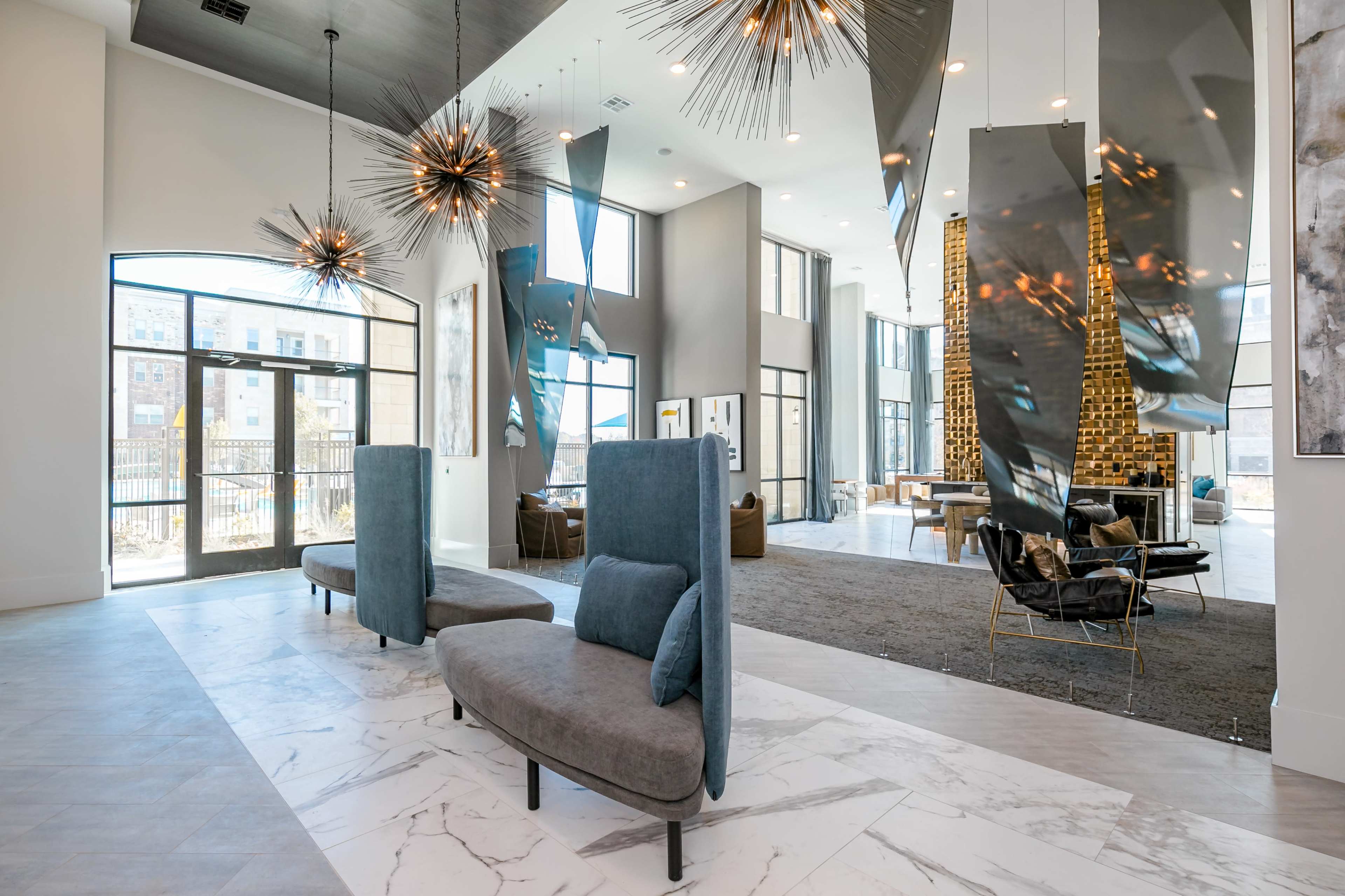 The image shows a modern lobby area featuring contemporary furniture, artistic light fixtures, and large windows that open to an outdoor space.