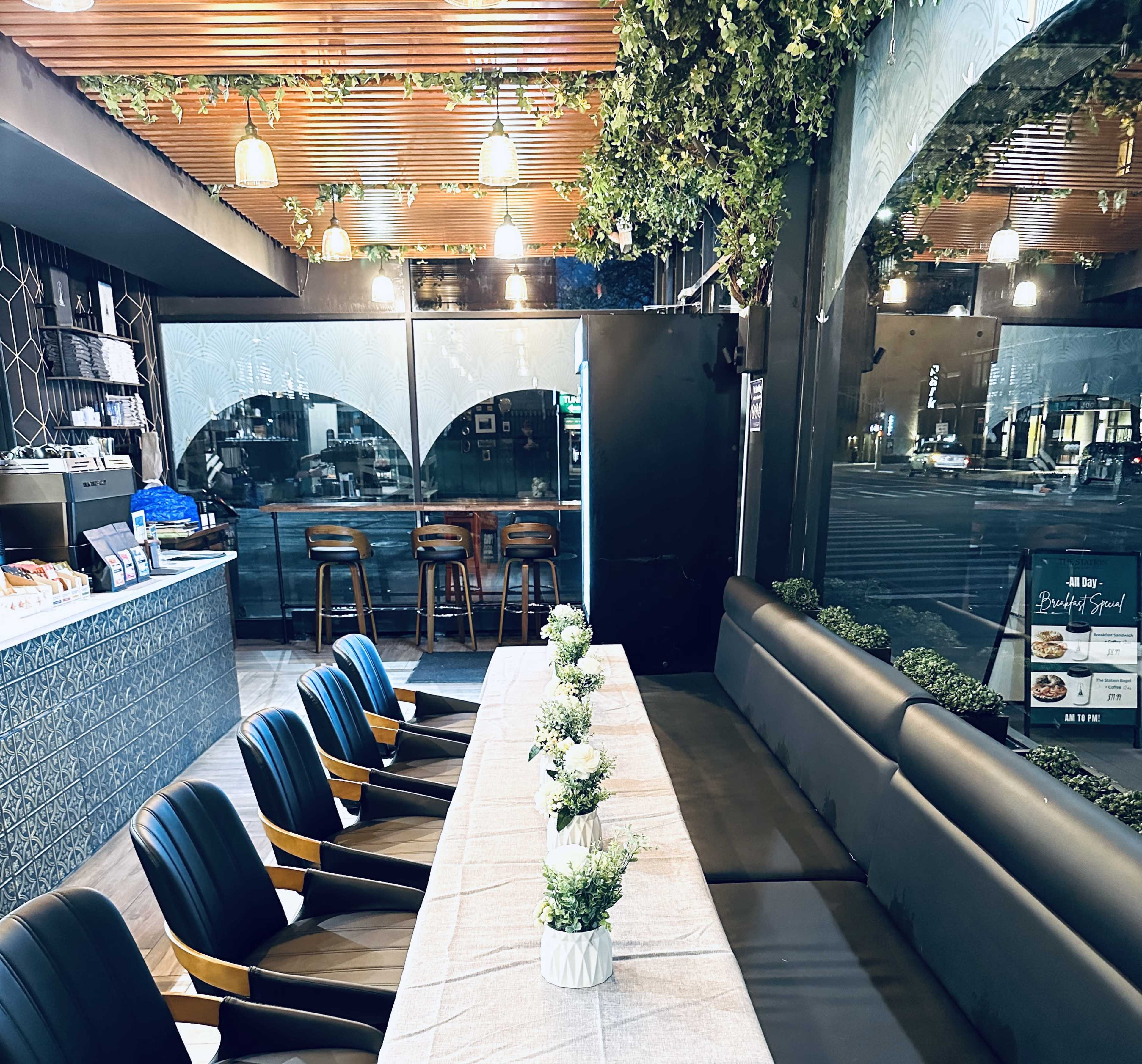 The image shows a modern café interior with a long table set for dining, surrounded by black chairs and adorned with small potted plants.