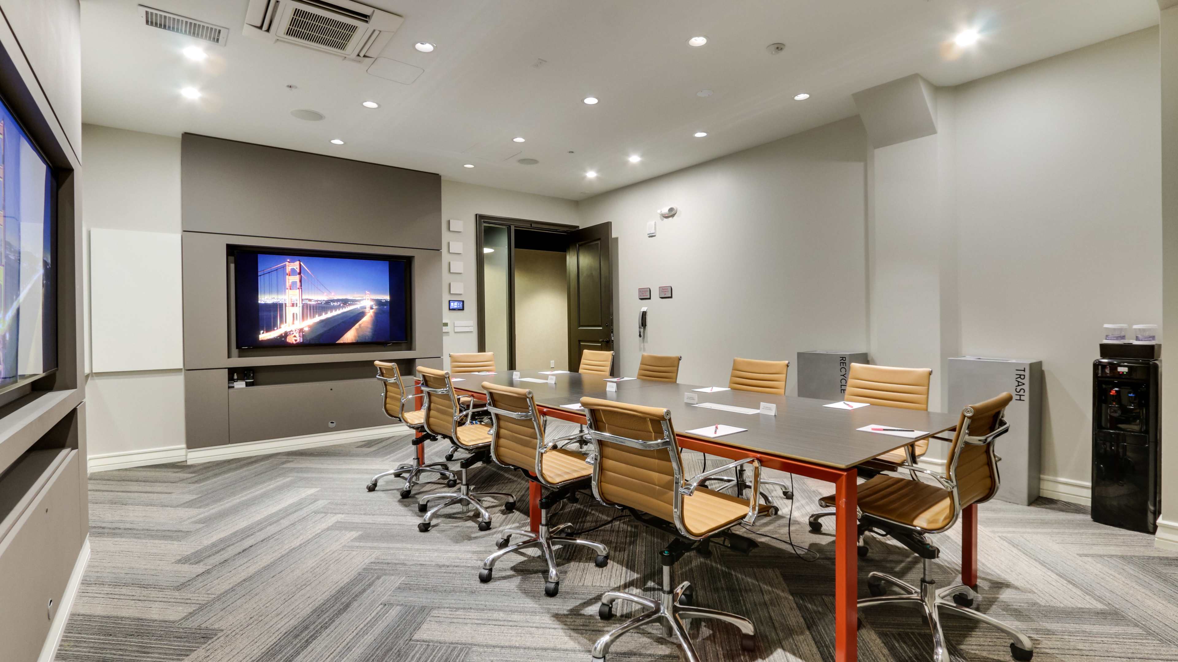 A modern conference room features a long table surrounded by orange-accented chairs, with a large screen displaying a cityscape.