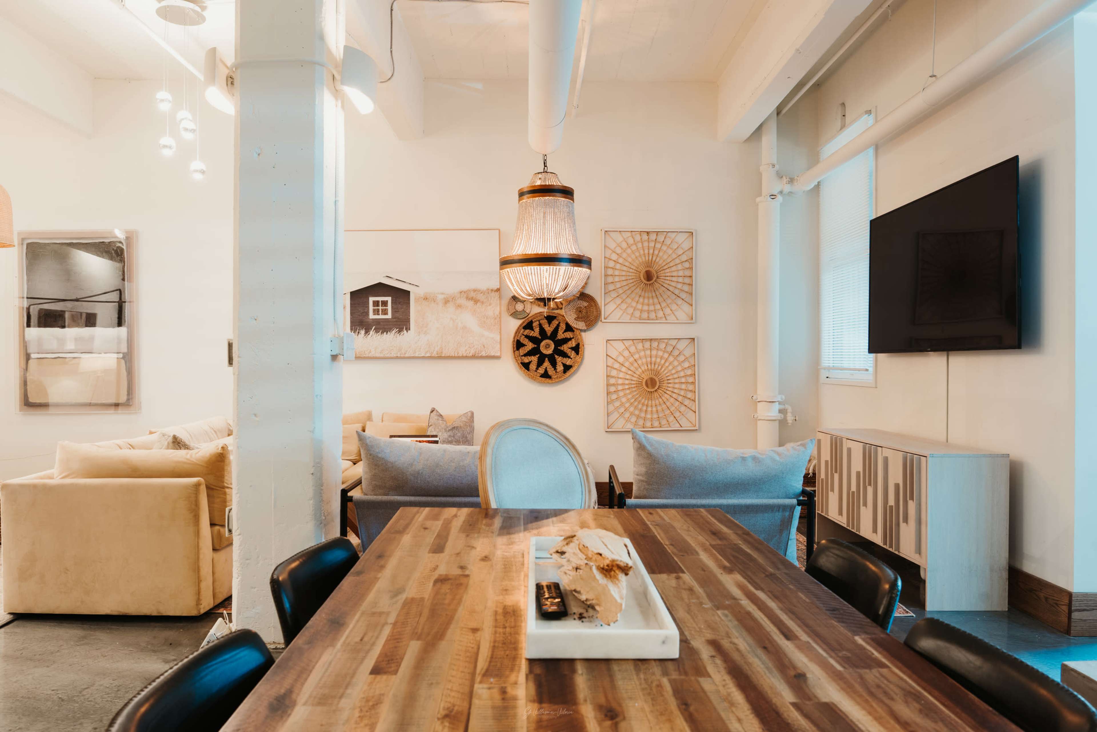 A dining table made of wood is centered in a modern, light-filled space decorated with wall art and featuring comfortable seating arrangements.