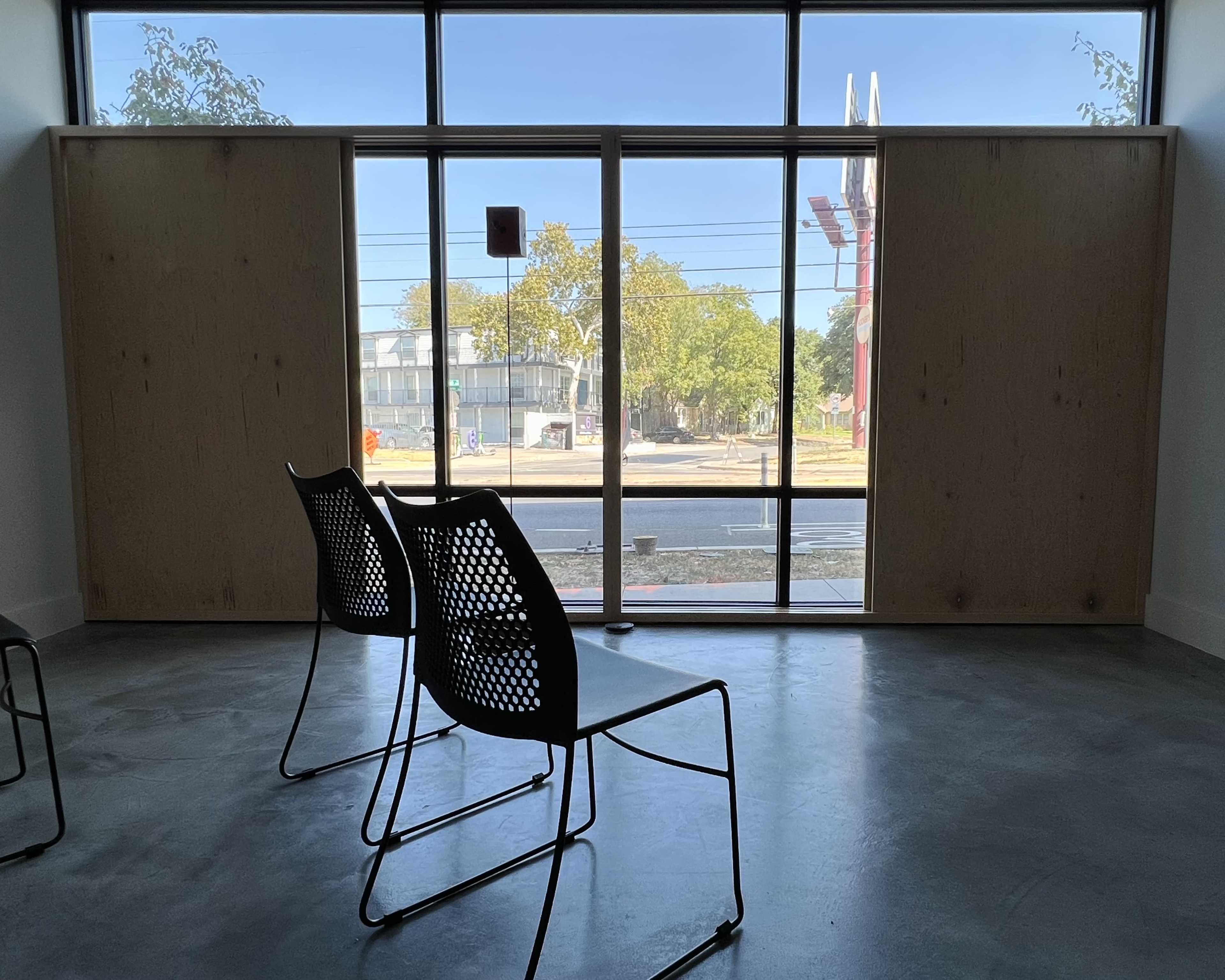The image shows two black wire chairs facing a large window with wooden panels on either side, revealing an outdoor scene with trees and a street.