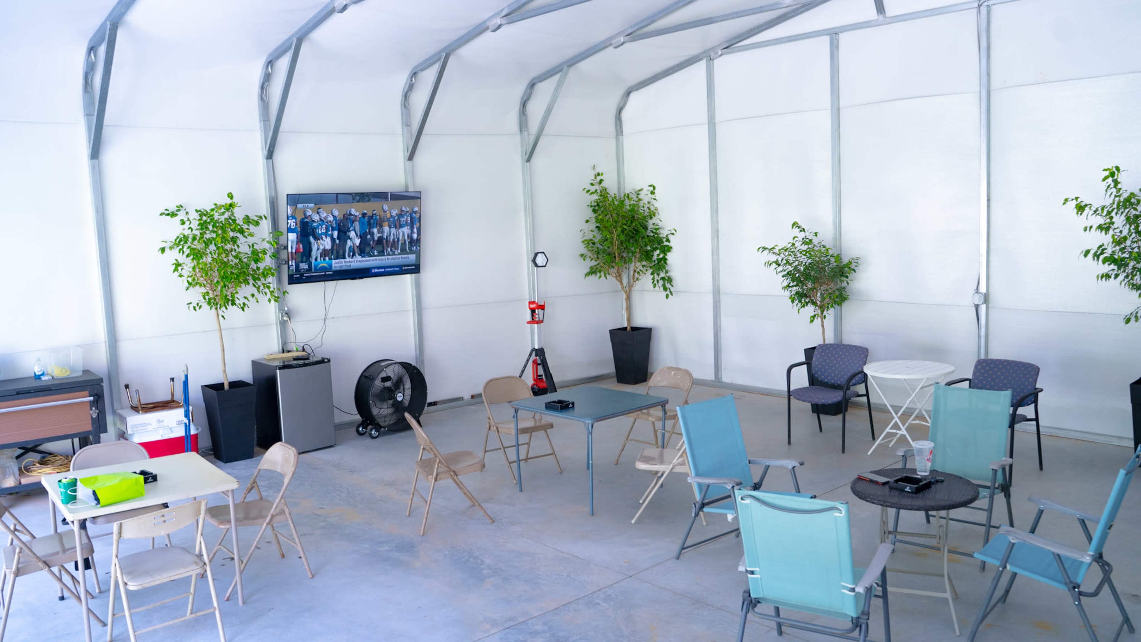 The interior of a spacious, white-walled structure is furnished with several tables and chairs, a television mounted on the wall, and two potted plants.