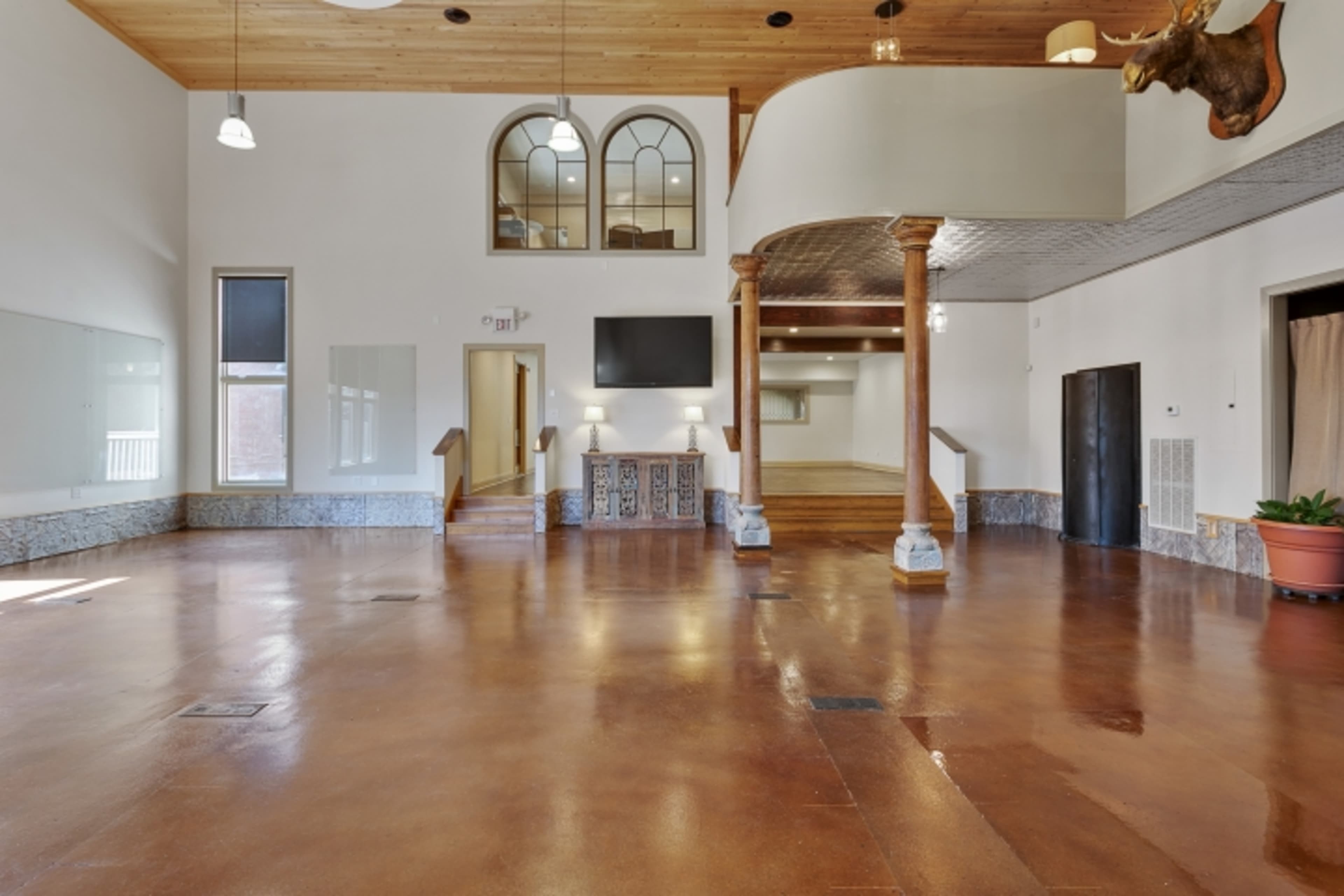 The image shows an empty interior space featuring a polished floor, wooden ceilings, decorative pillars, and large windows.