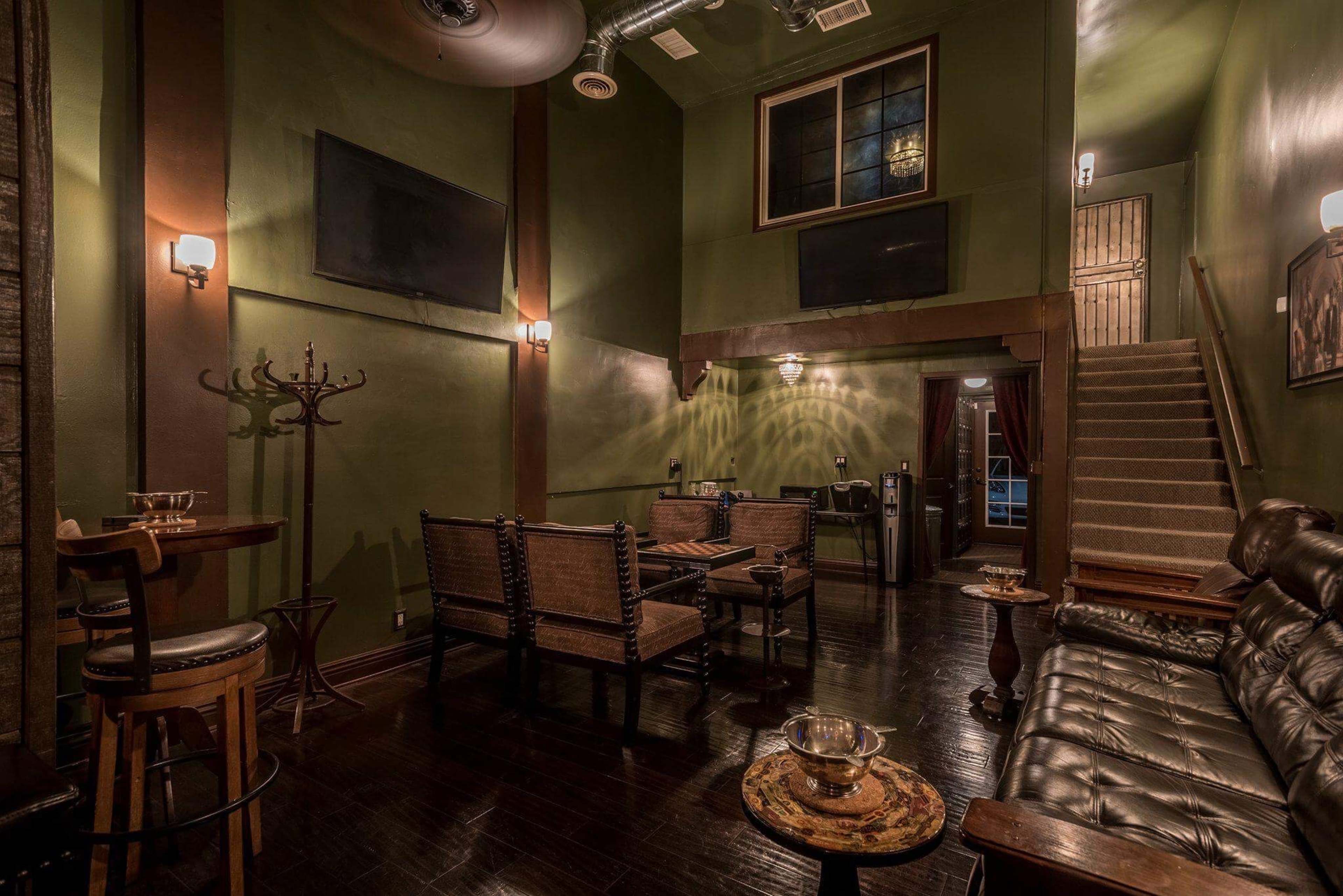 A dimly lit lounge with dark green walls, wooden floors, a leather sofa, wicker seating, and a staircase leading to an upper level.