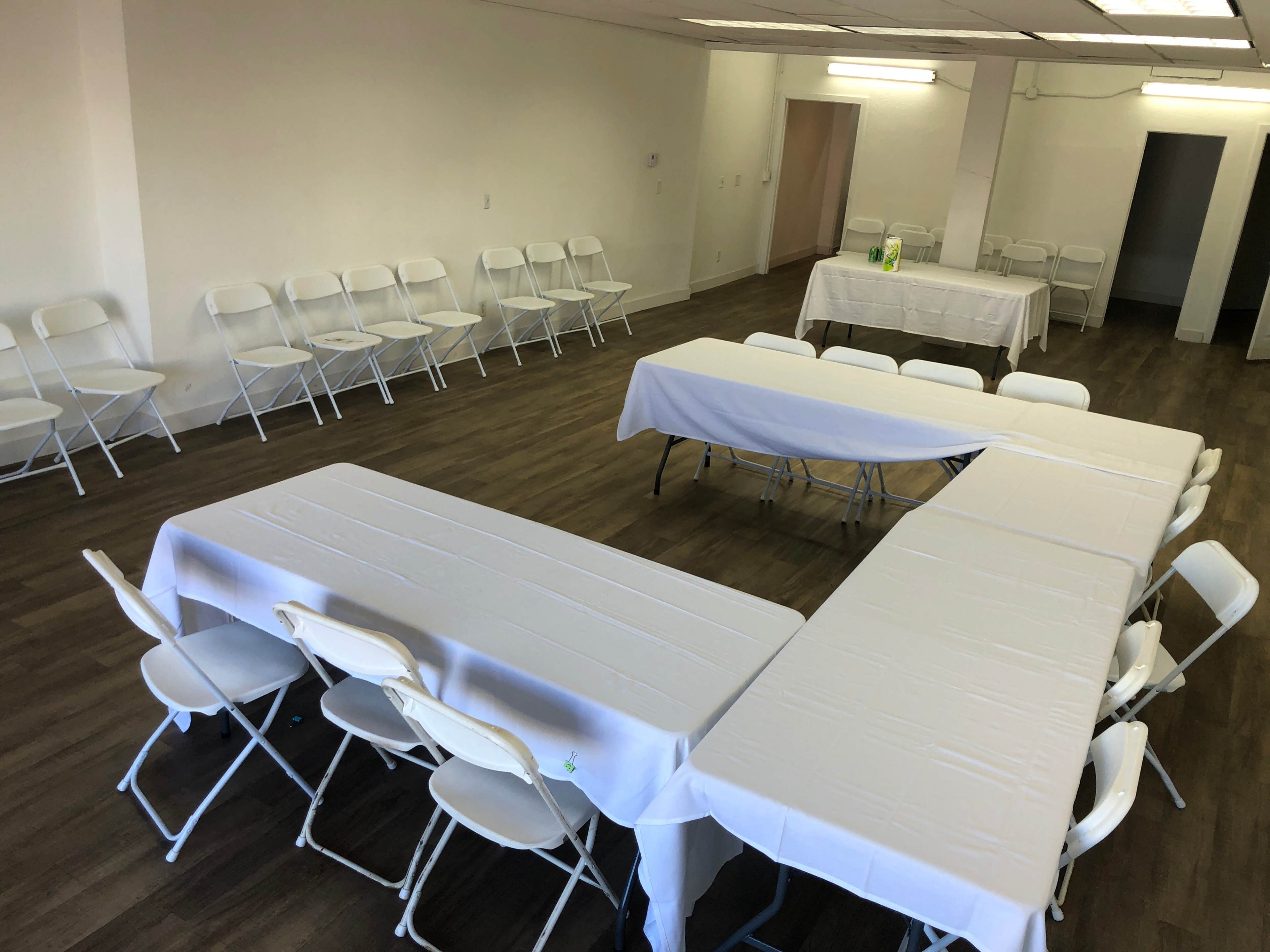 A large, empty room features several tables arranged in a U-shape with white tablecloths, surrounded by white folding chairs.