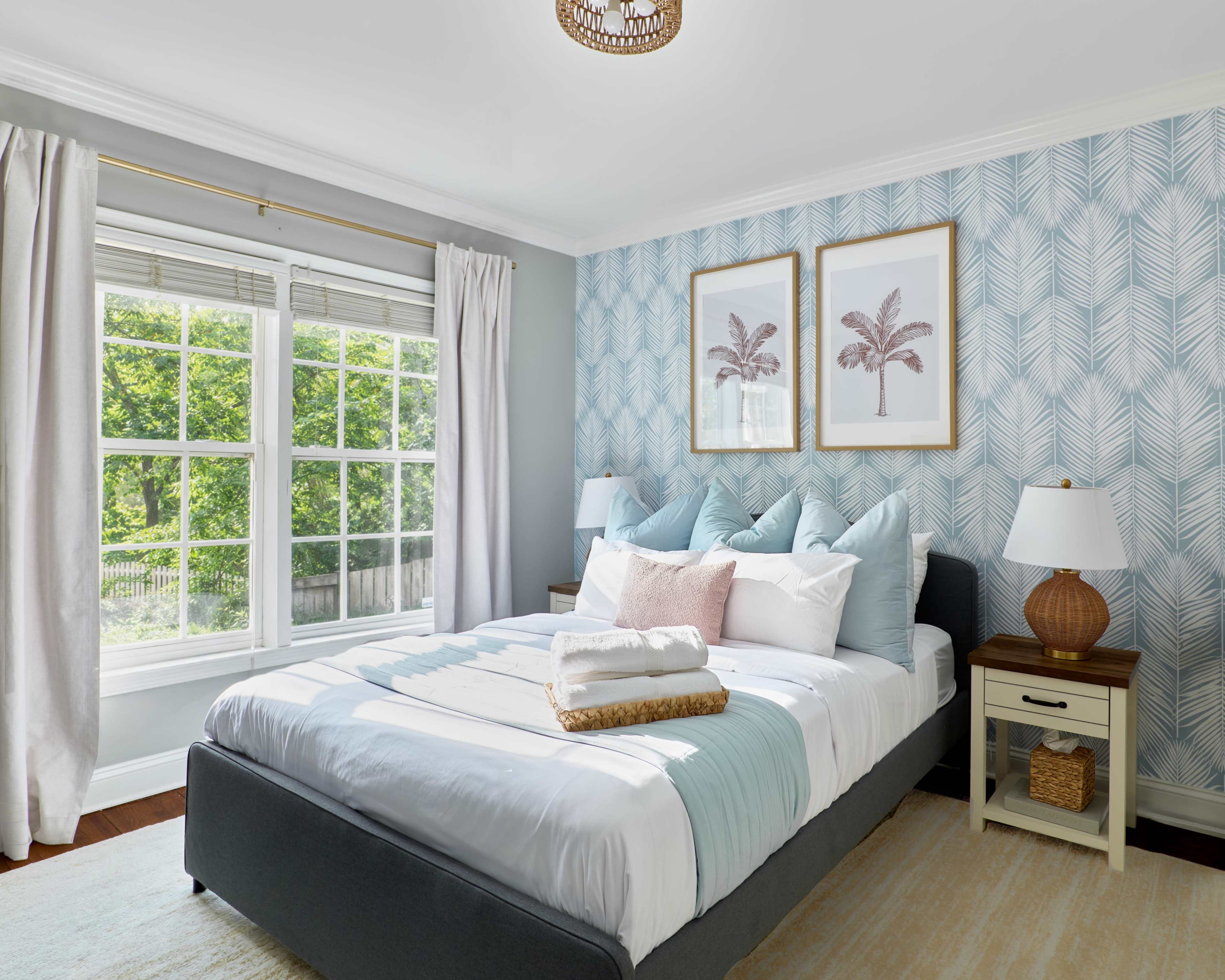 A neatly arranged bedroom features a large bed with decorative pillows, two framed palm prints on the wall, and a window revealing greenery outside.