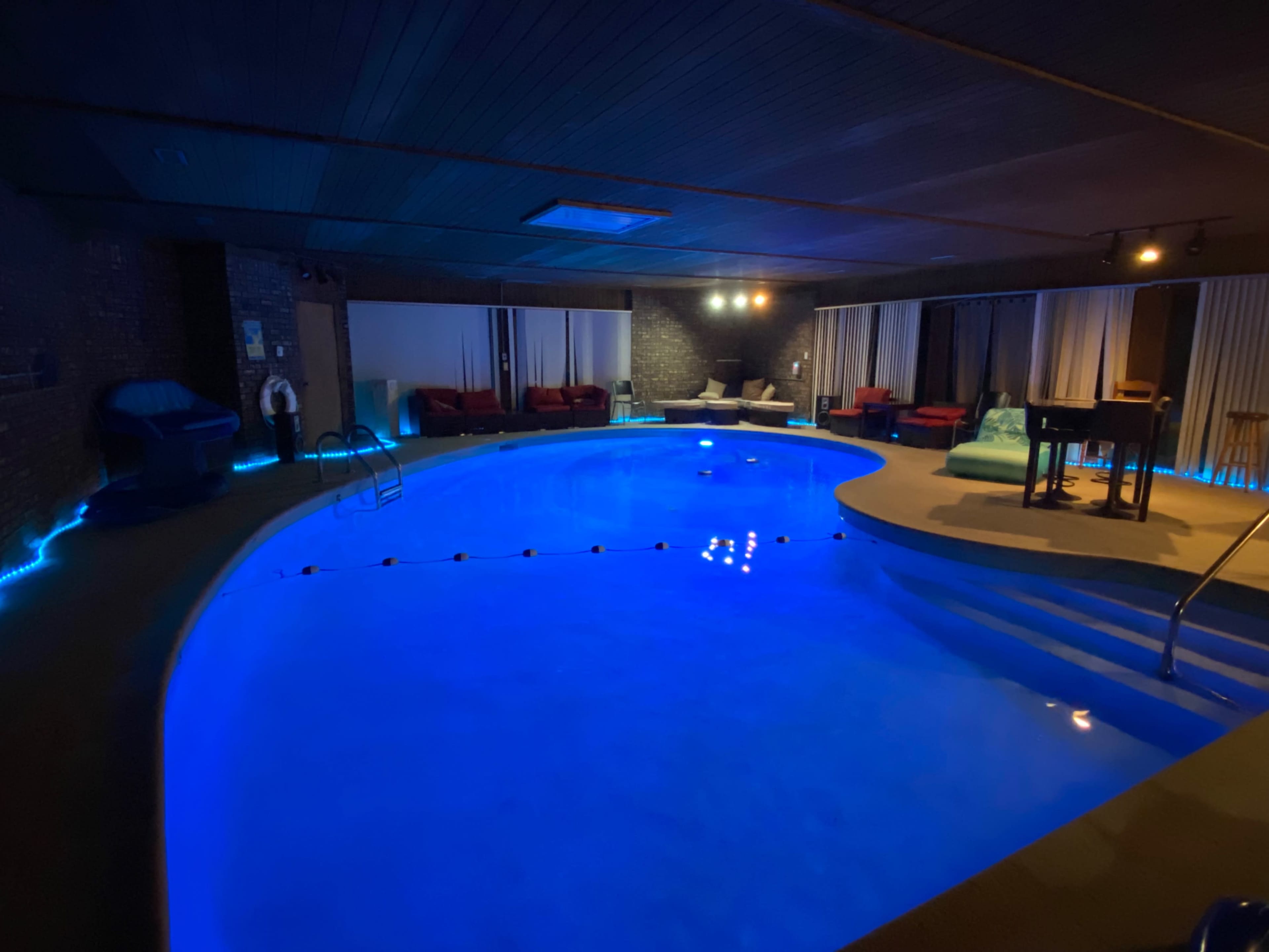 An indoor pool area with a blue-lit pool, surrounded by lounge furniture and curtains.