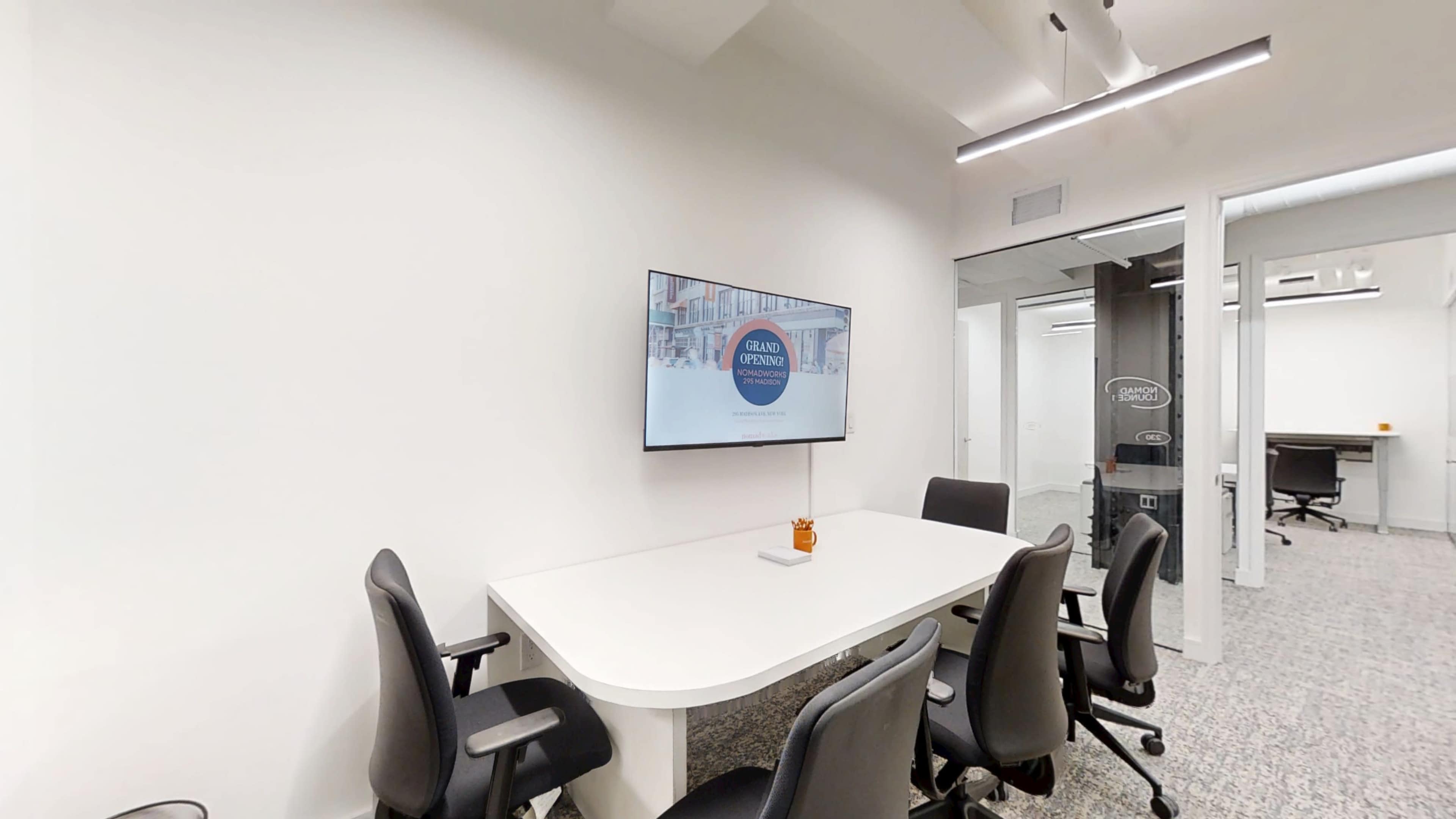 The image shows a modern meeting room with a long white table, several black office chairs, and a wall-mounted display screen.