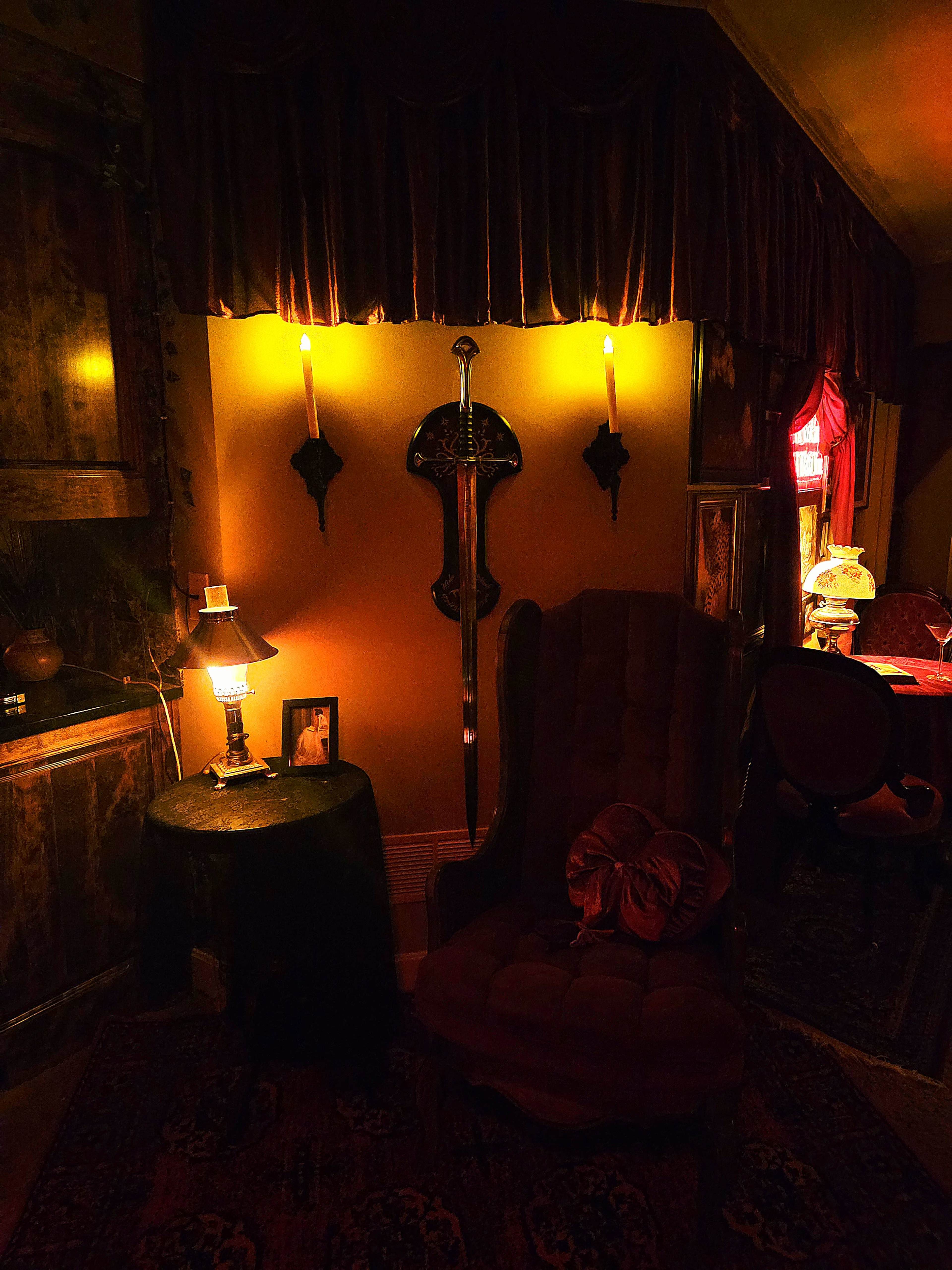 The image depicts a dimly lit room with vintage decor, featuring a plush armchair, a small table with a lamp, and wall sconces flanking a decorative shield.