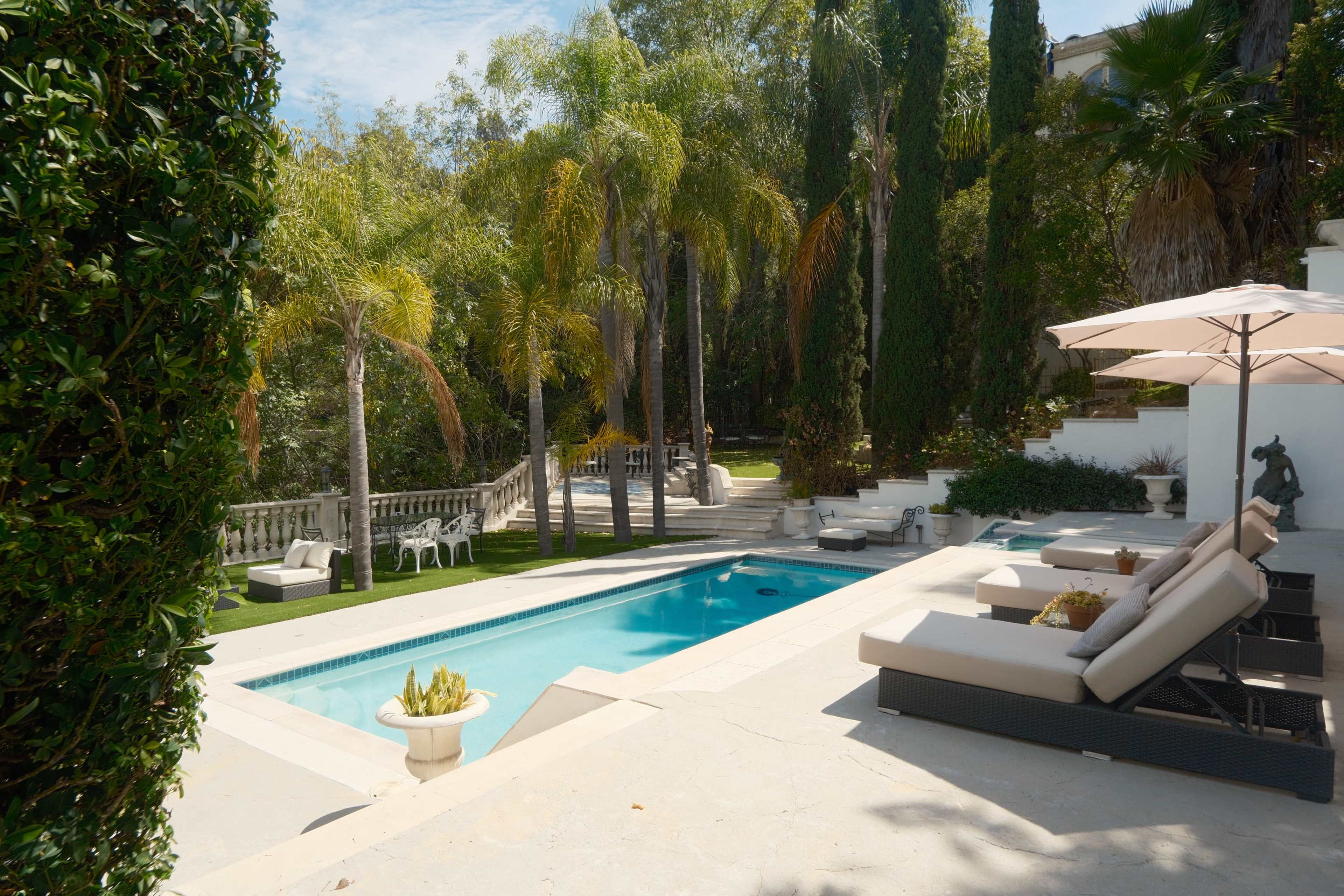 The image depicts a serene outdoor pool area surrounded by palm trees, lounge chairs, and stone pathways, with a view of a landscaped garden in the background.