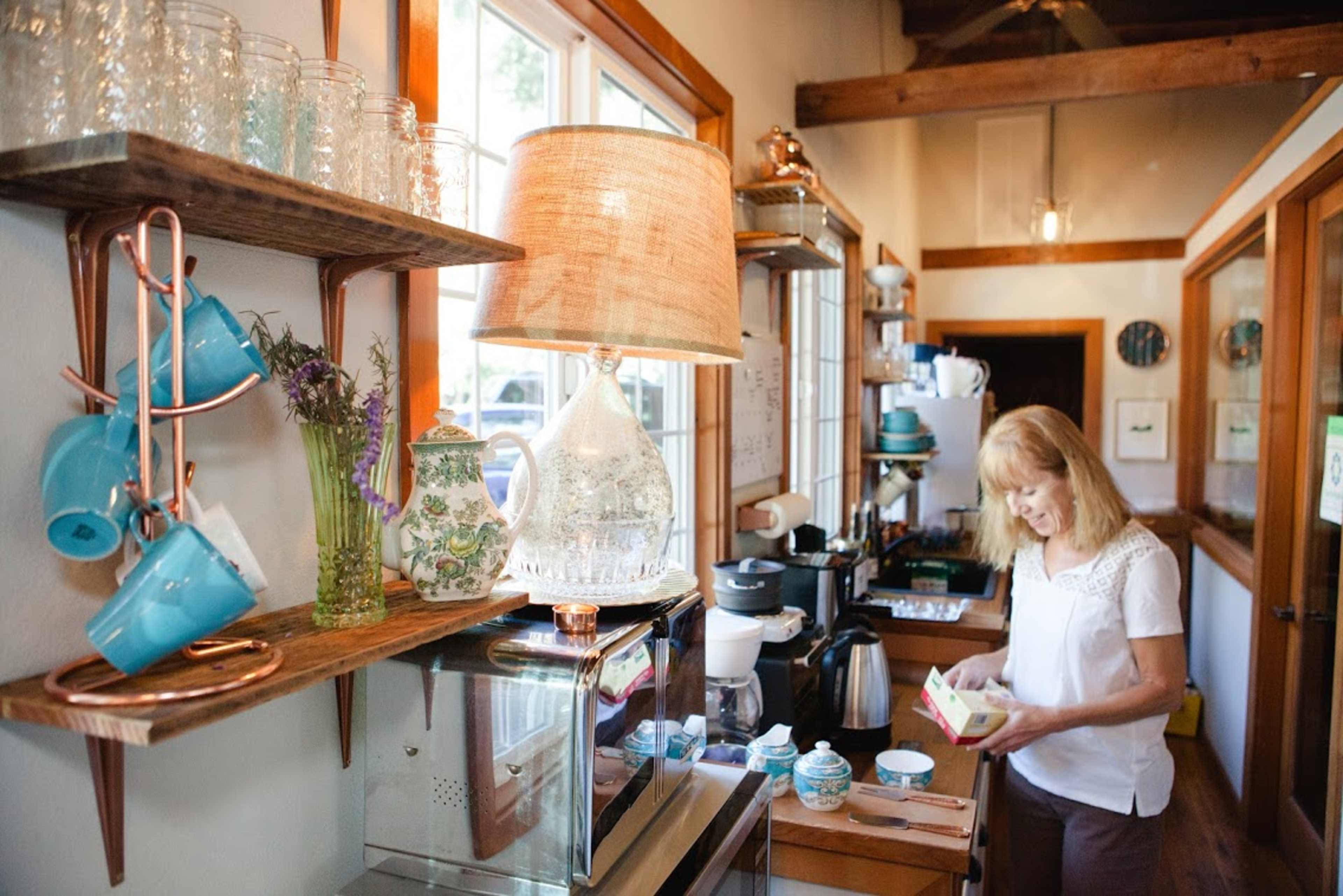 A woman organizes items on a countertop in a cozy kitchen with shelves displaying colorful mugs and kitchenware.