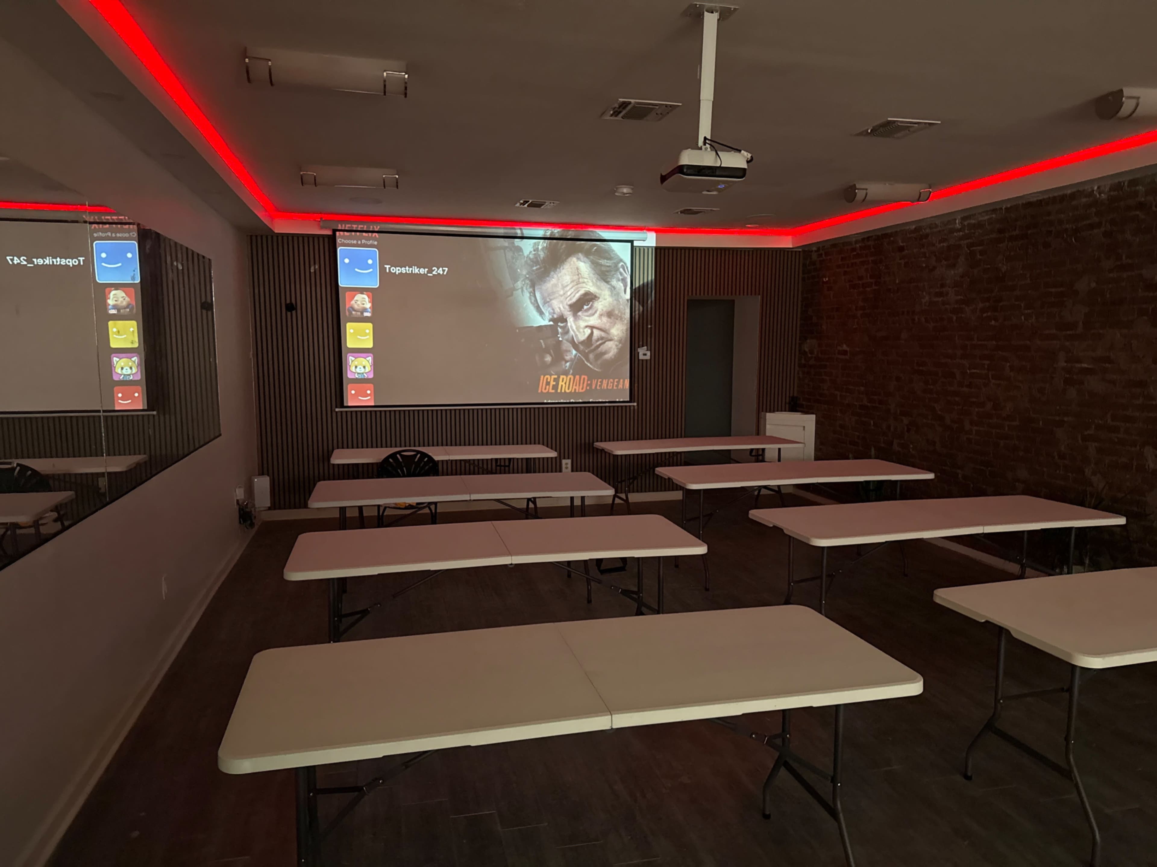 The image shows a sparsely furnished room with several tables arranged for an event, featuring red lighting and a projection of a movie on the wall.