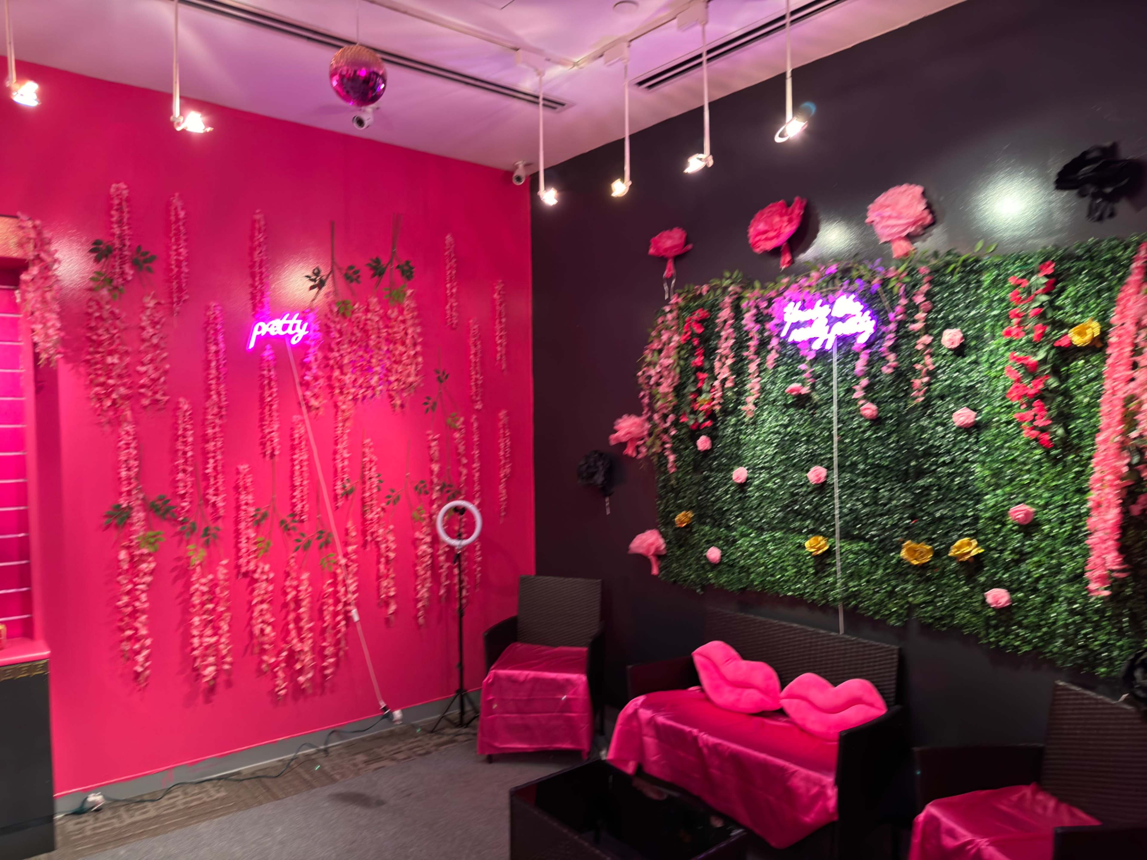 A brightly colored room with pink walls adorned with artificial flowers and neon signs, featuring seating arrangements and decorative elements.
