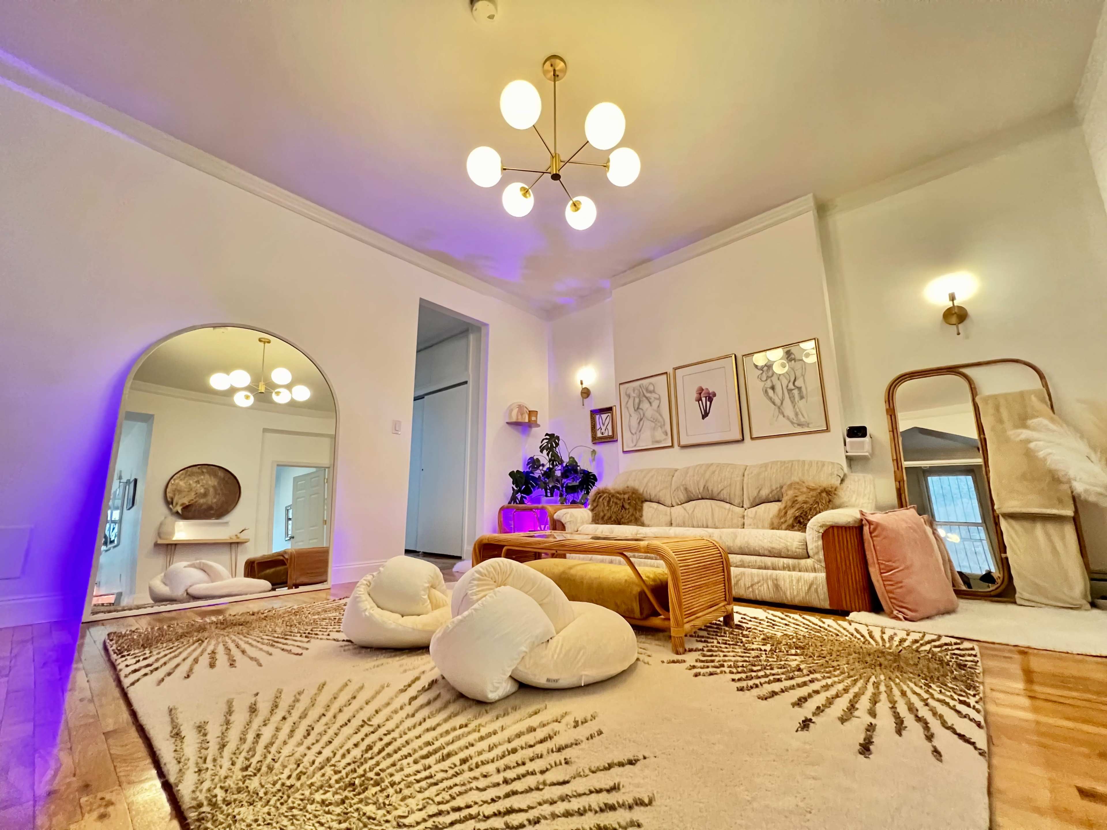 A well-decorated living room features a large beige sofa, a round mirror, and a decorative rug with gold patterns, illuminated by a modern light fixture.