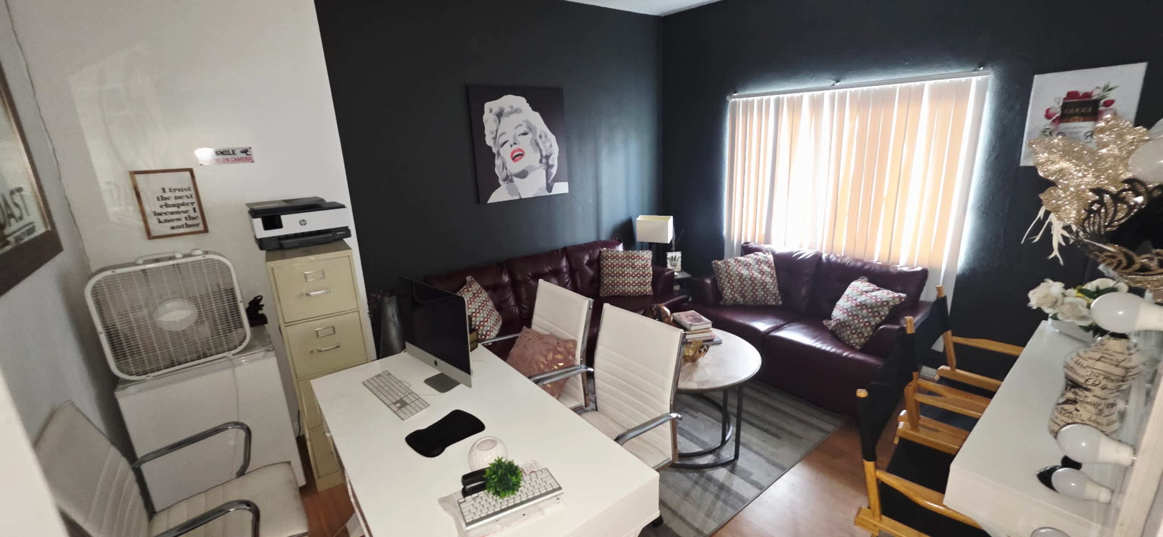 The image shows an office space featuring two sofas, a desk with chairs, a printer, and decorative elements against a dark wall.