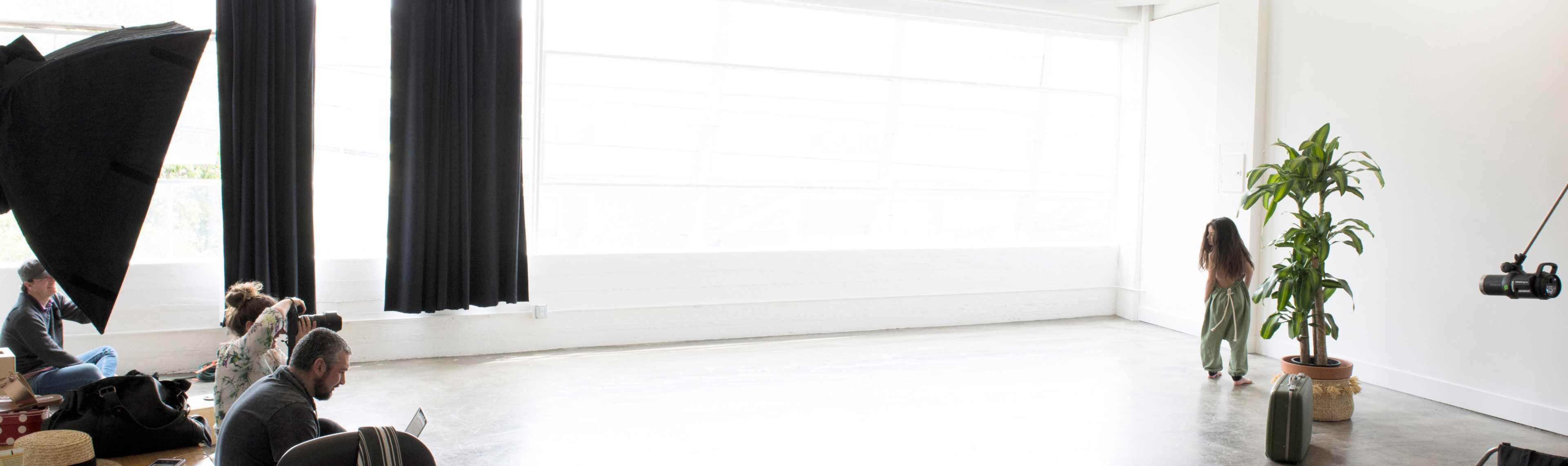 A child stands on a bare floor in a brightly lit studio space, while an audience sits on the left side, observing.
