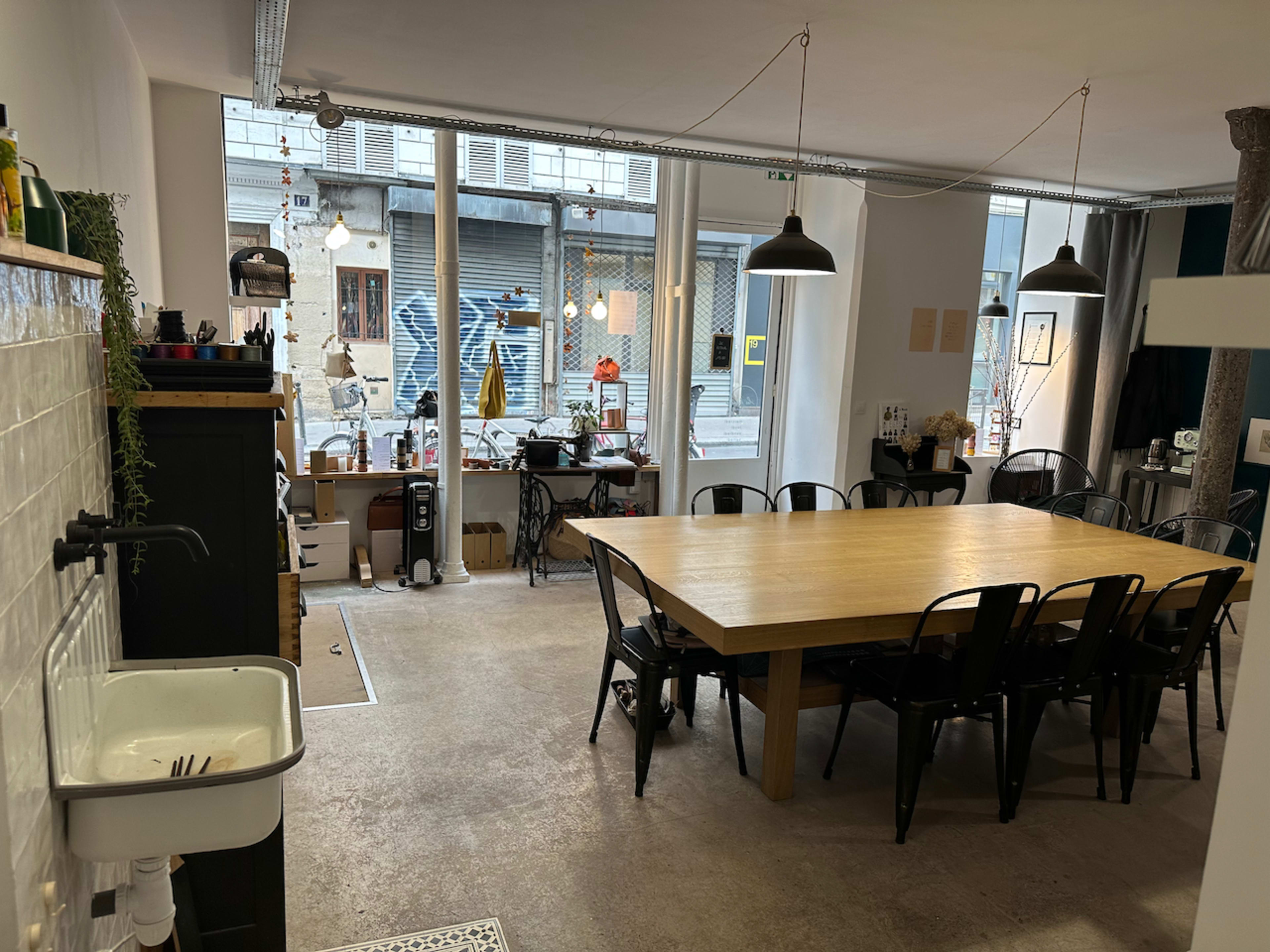 A spacious, modern kitchen area features a large wooden table surrounded by black chairs, with large windows showing an urban street outside.