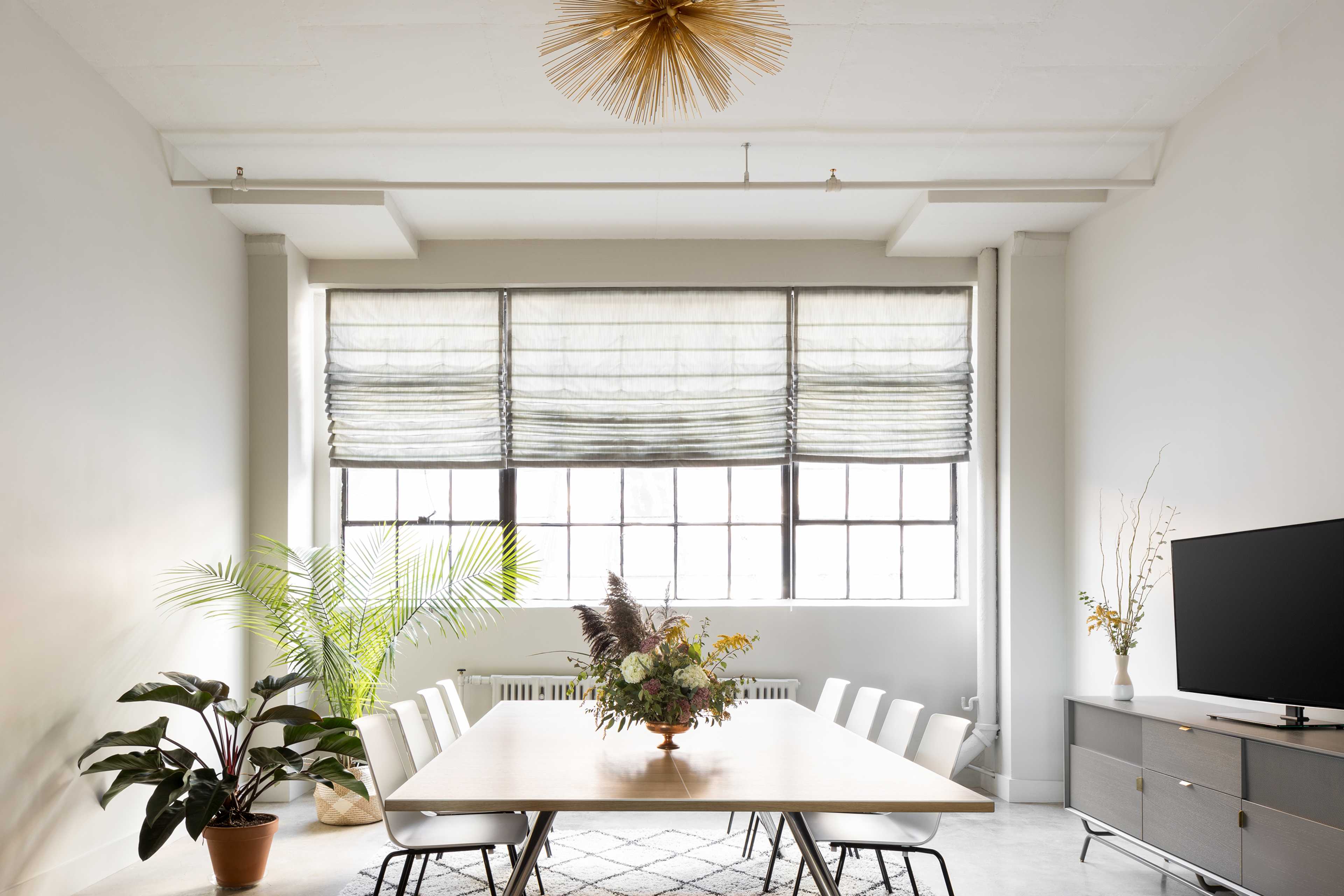 A modern dining room features a large table surrounded by white chairs, a decorative plant in the corner, and a television on a low cabinet beneath large windows with textured shades.